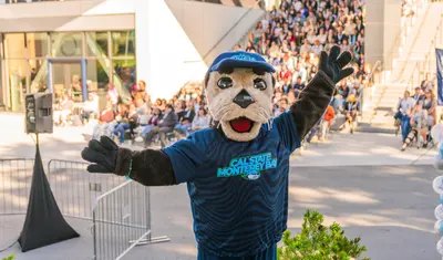Person in a sea otter mascot costume wearing a Cal State Monterey Bay shirt, waving to a crowd.