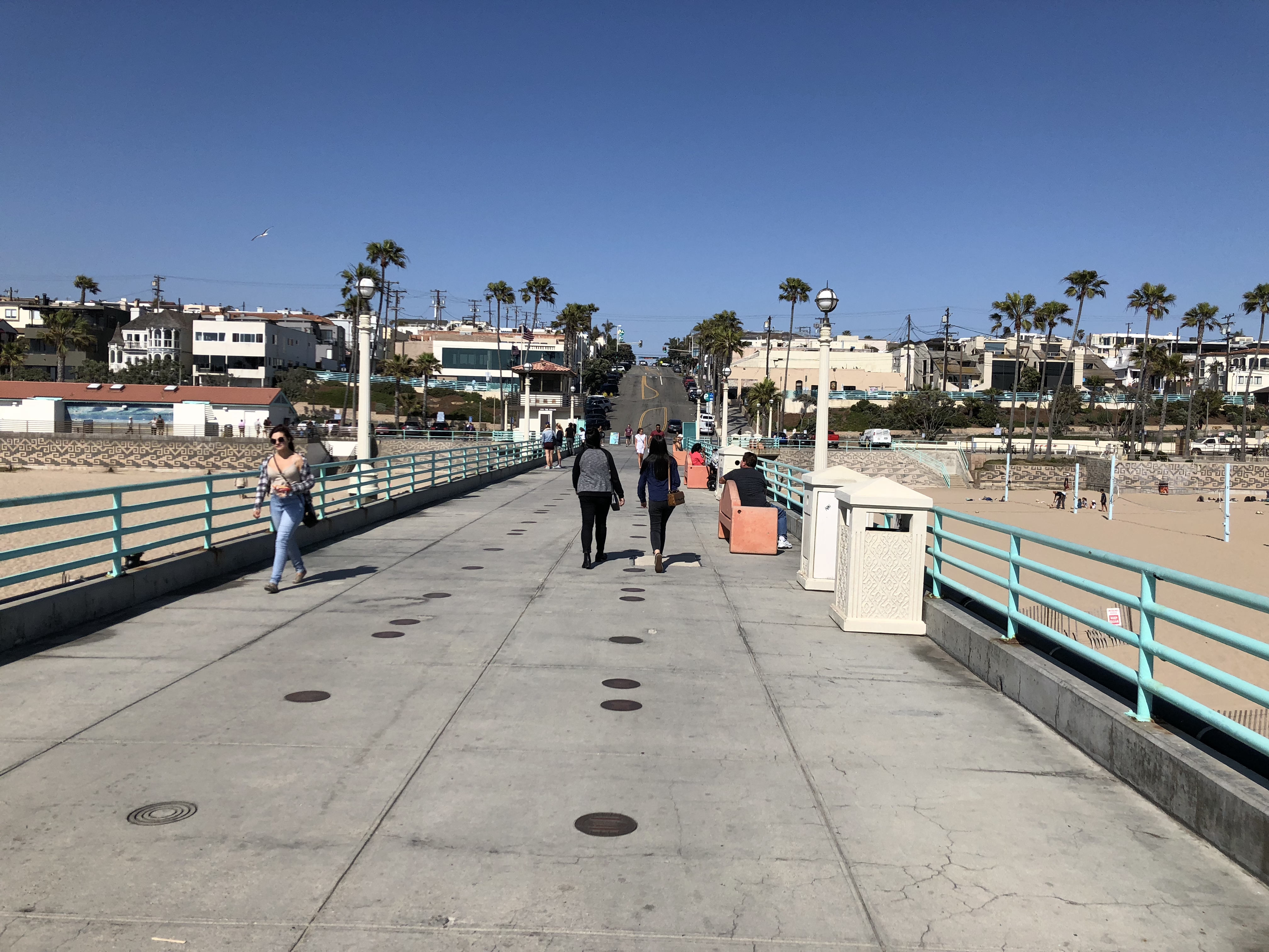 Pedestrians walk along a wide pier with teal railings toward a palm-tree-lined beach.