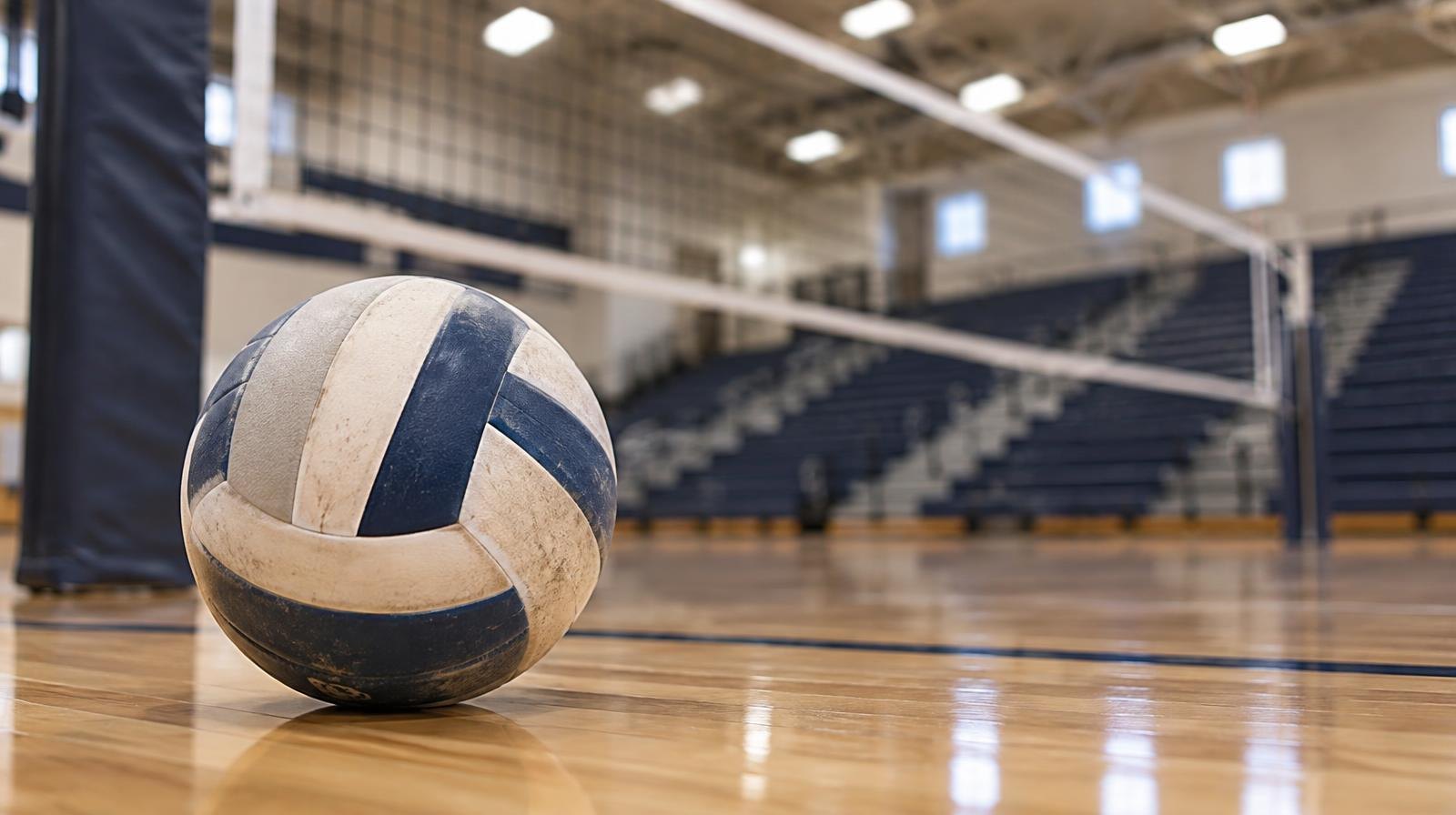 A volleyball resting on a hardwood gym floor near a net post, bleachers blurred behind it.