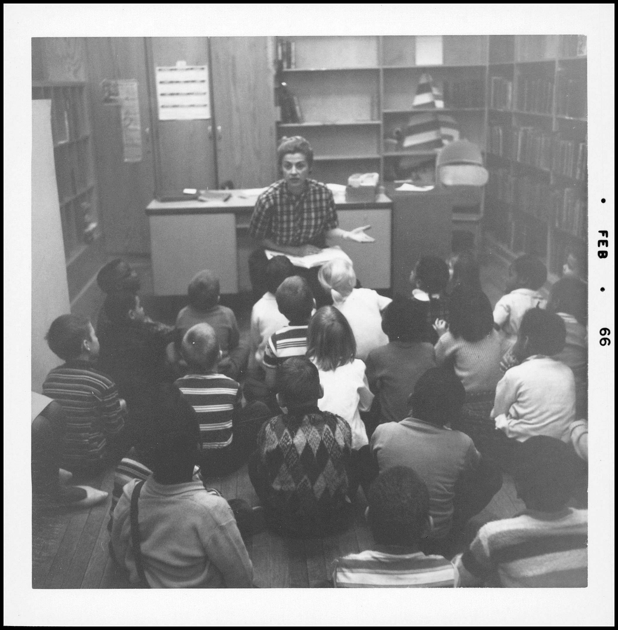 A librarian reads aloud to children seated on the floor in a school library.