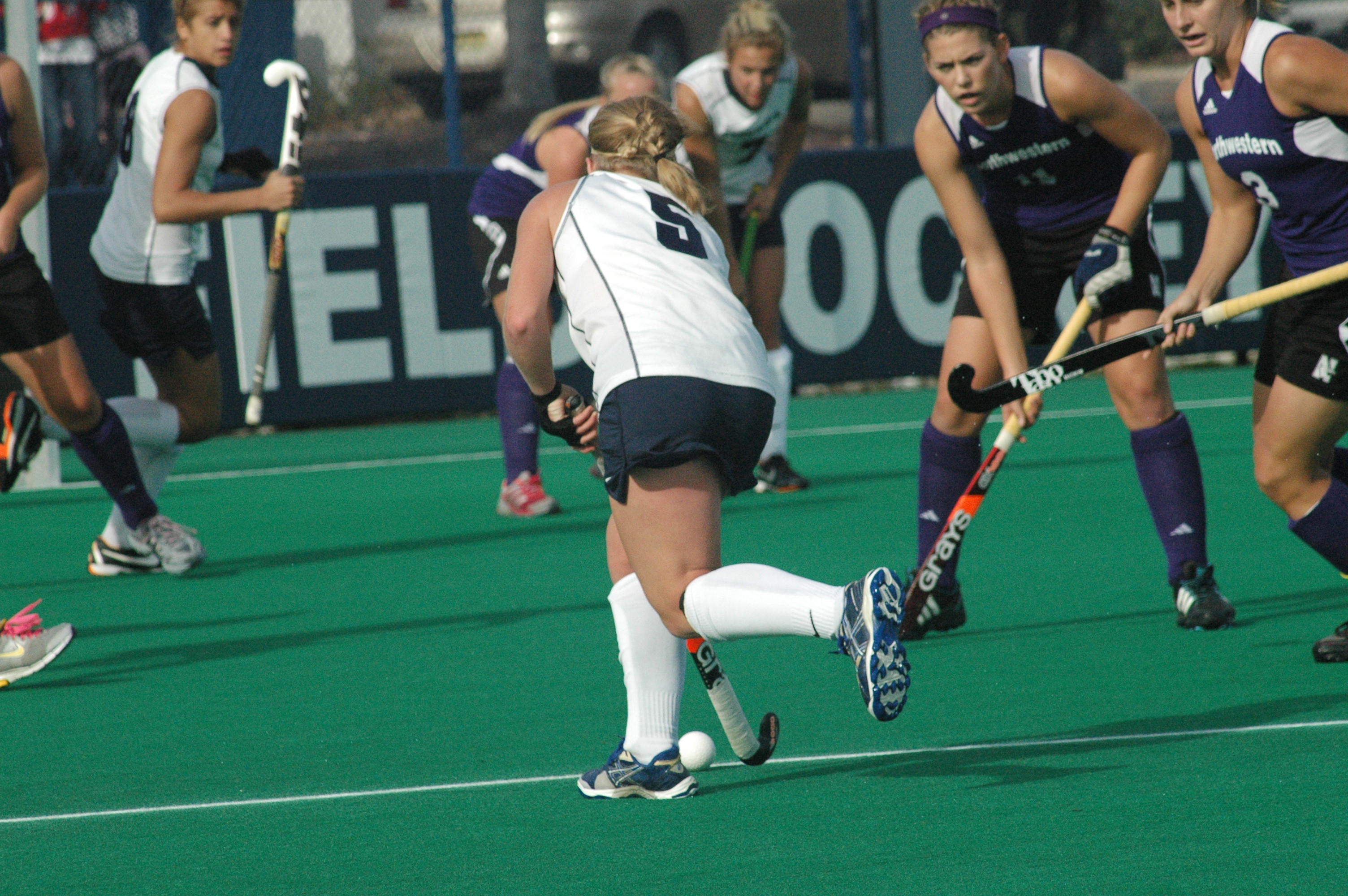 Female field hockey player in white dribbling the ball while purple-uniformed defenders close in on a turf field.