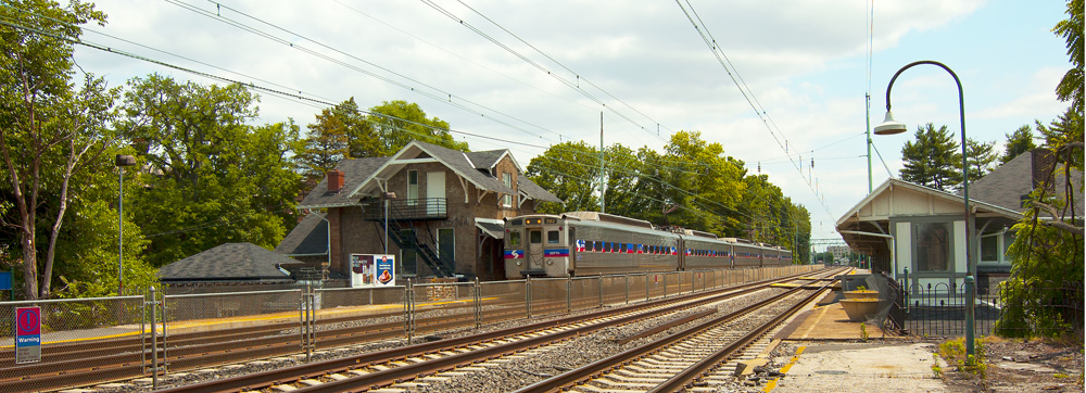 A commuter train stopped at a suburban rail station platform with multiple tracks and overhead wires.