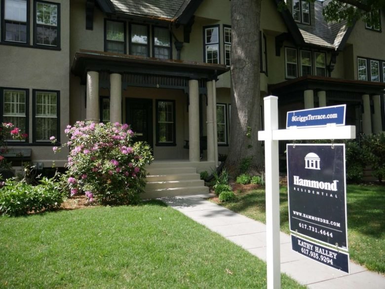 A large residential home with a real estate for-sale sign, covered porch, and flowering bushes in the front yard.