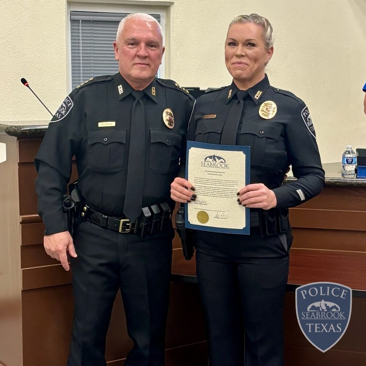 Two uniformed police officers stand together, one holding a certificate with a gold seal.