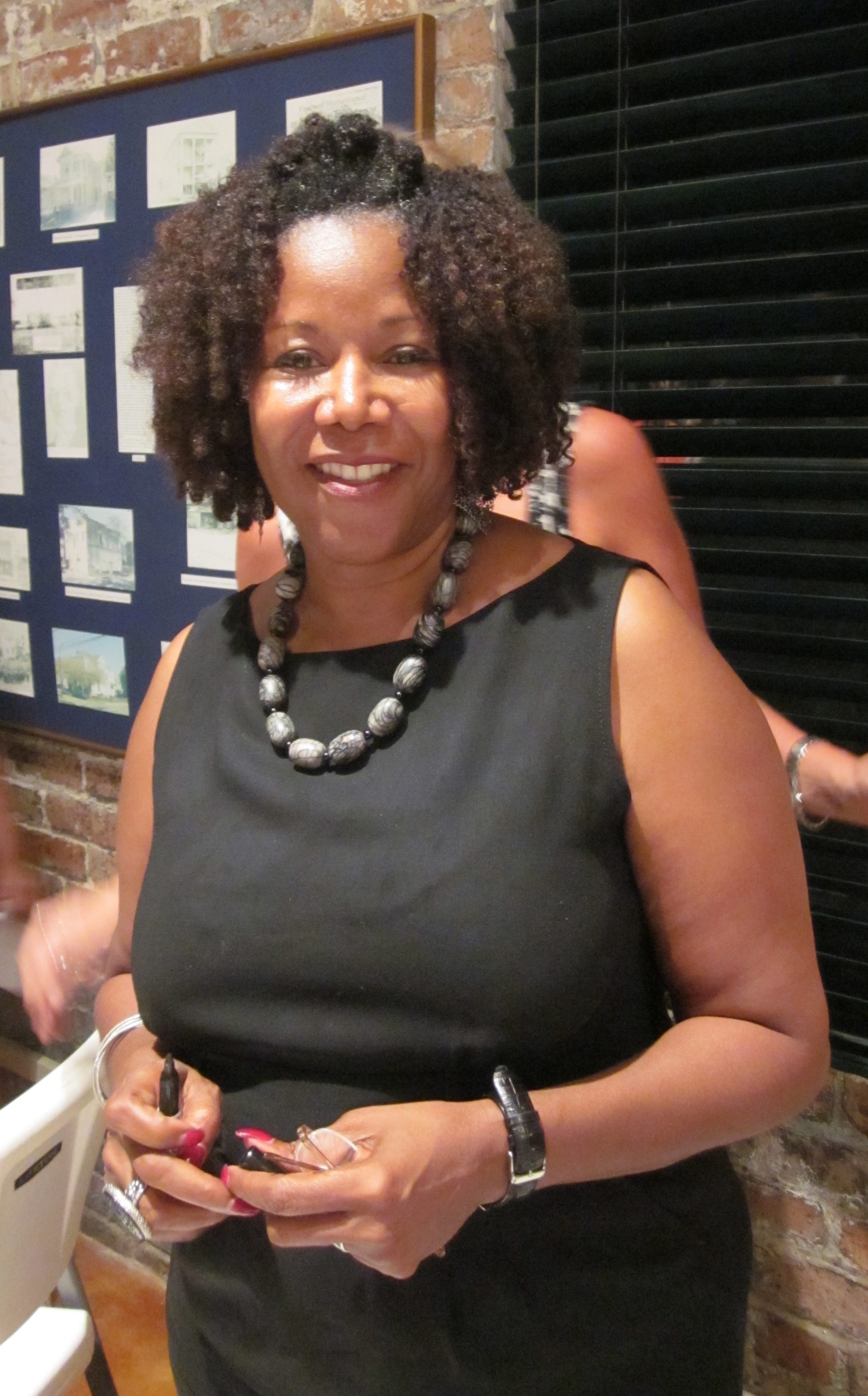 Woman in a black dress smiling and holding a pen indoors, standing before a photo display.