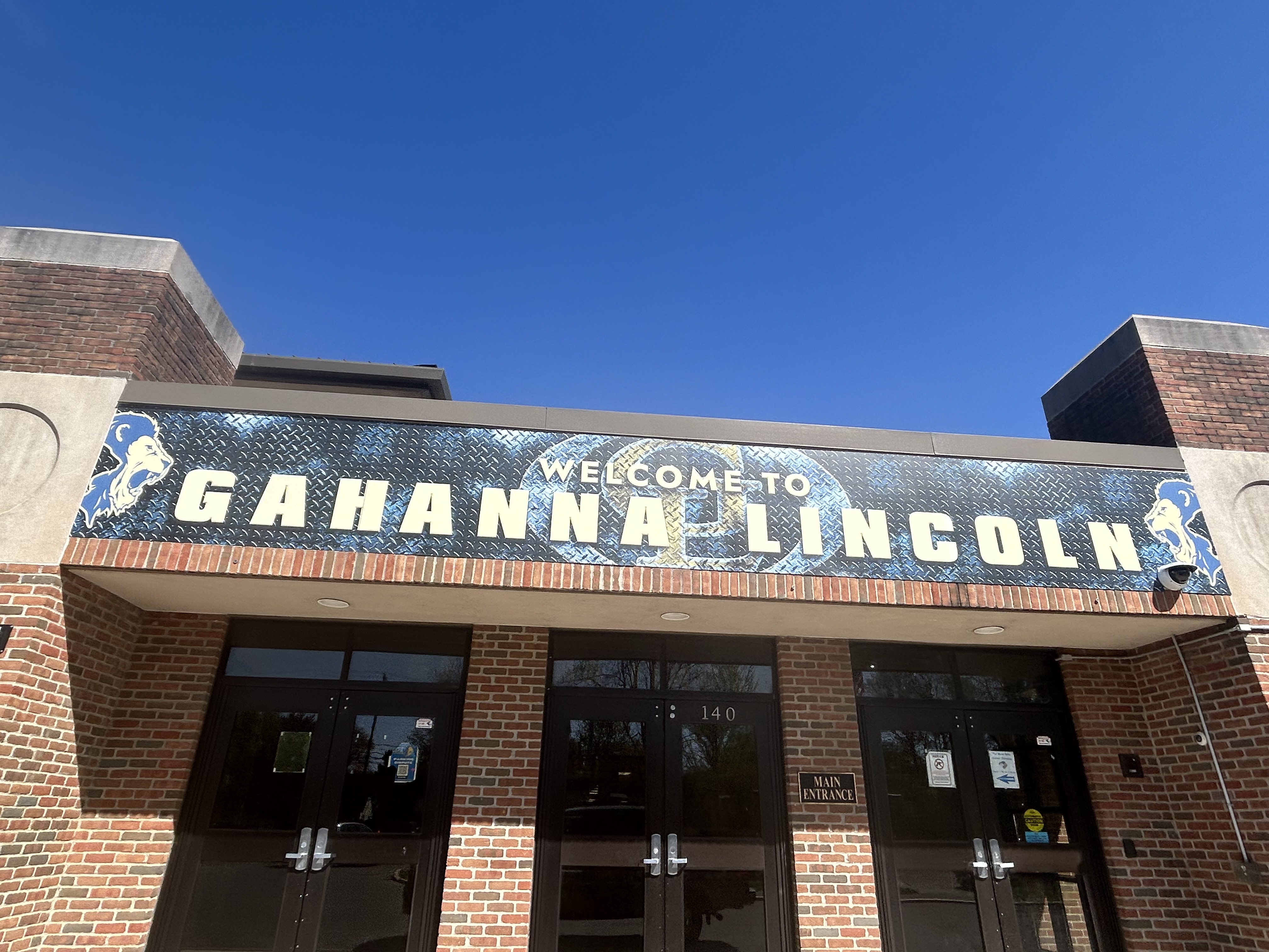 Blue and silver welcome sign above the brick main entrance of a high school building.