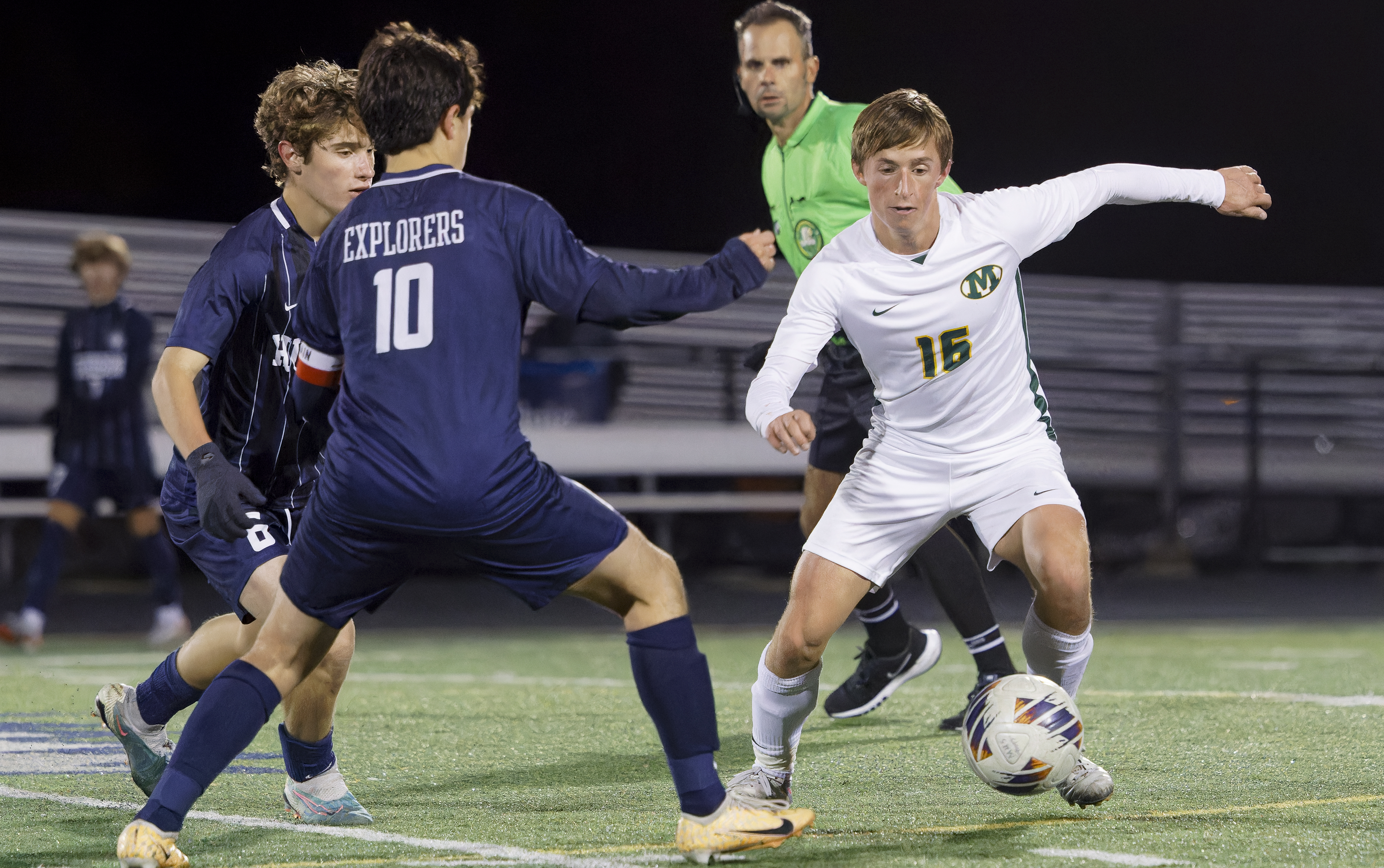 Three high school soccer players competing for the ball on a lit turf field at night.