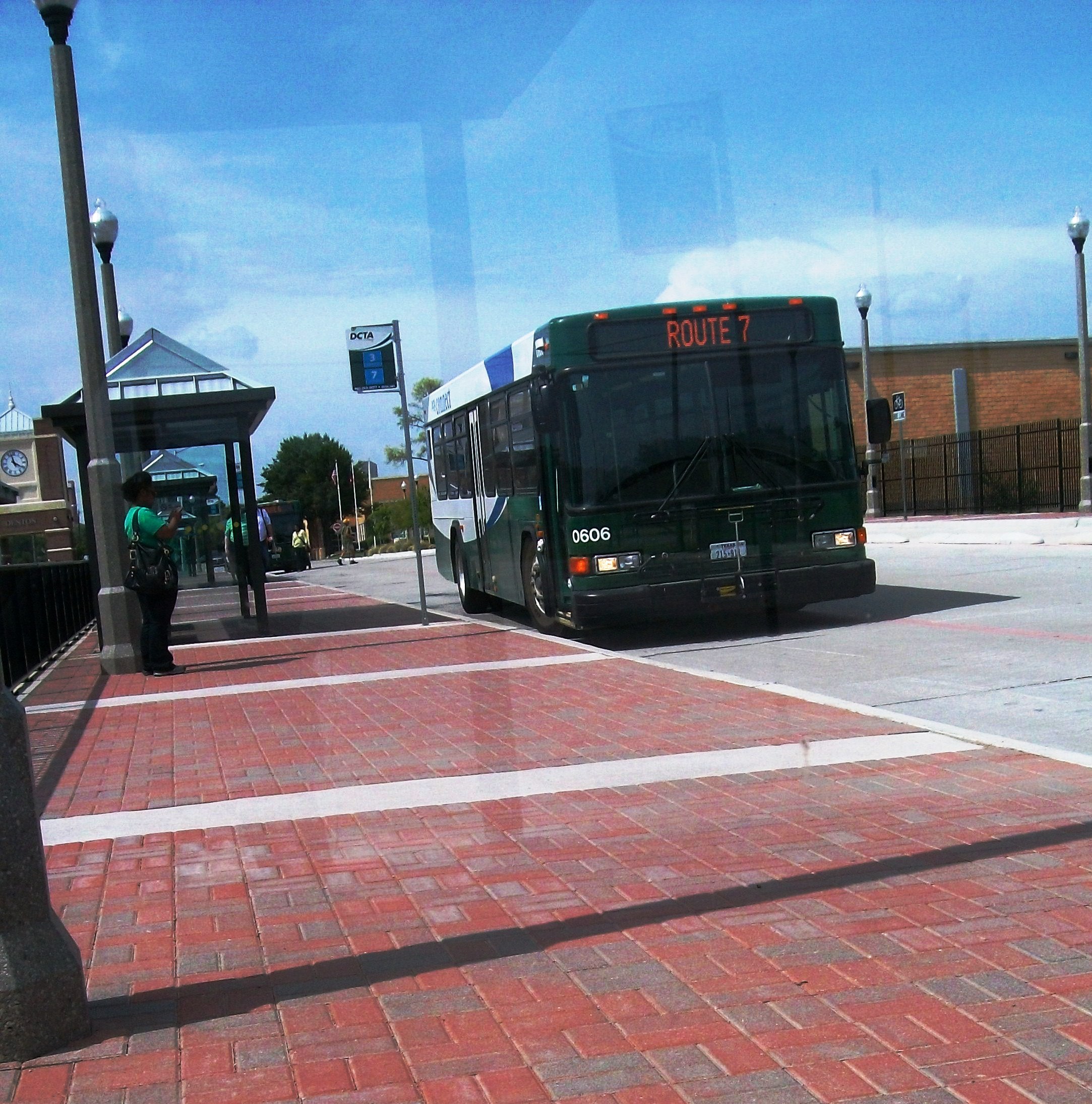 A green DCTA bus stopped at a brick-paved transit center with a covered shelter.