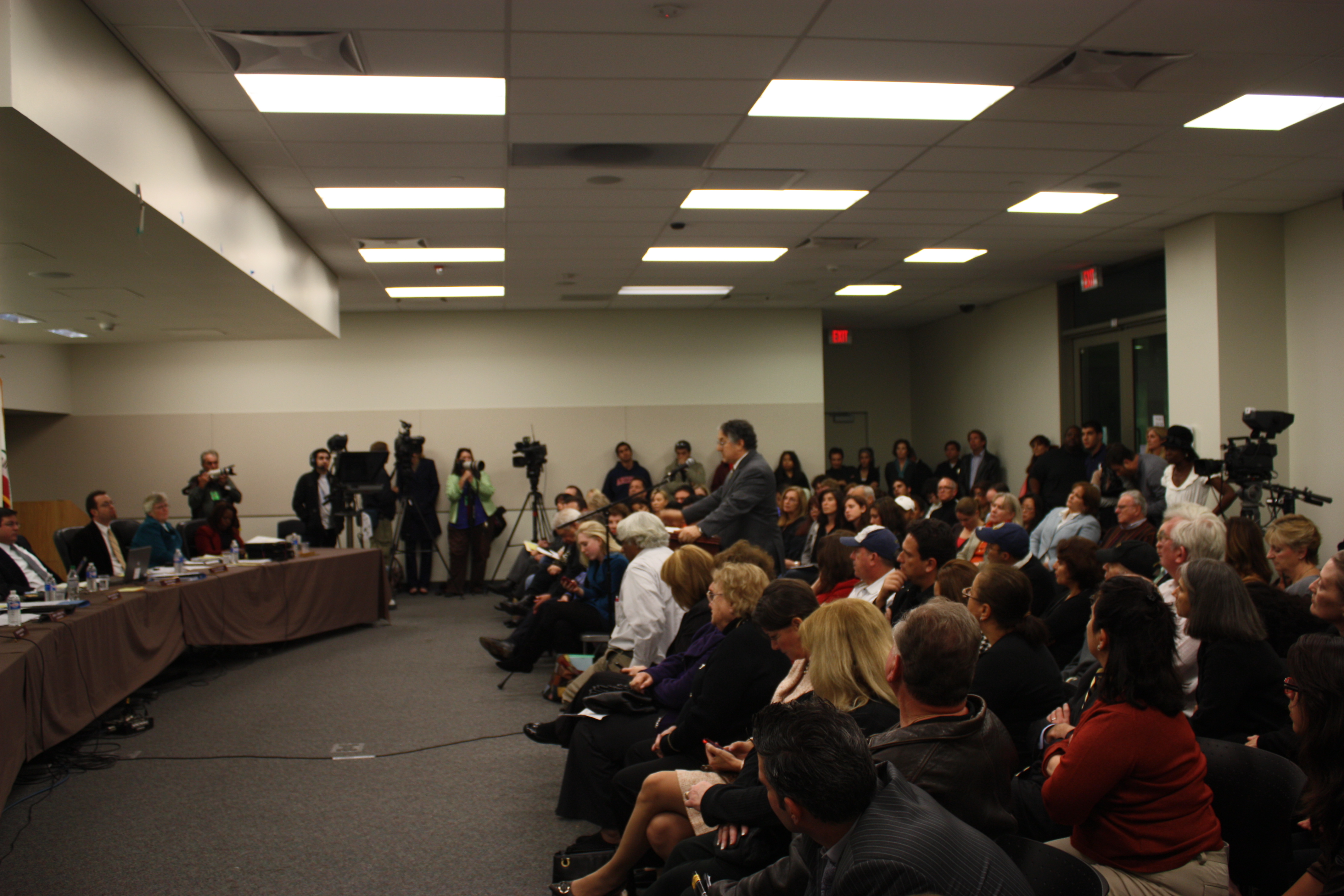 Crowded school board meeting room with officials seated at a front panel table.