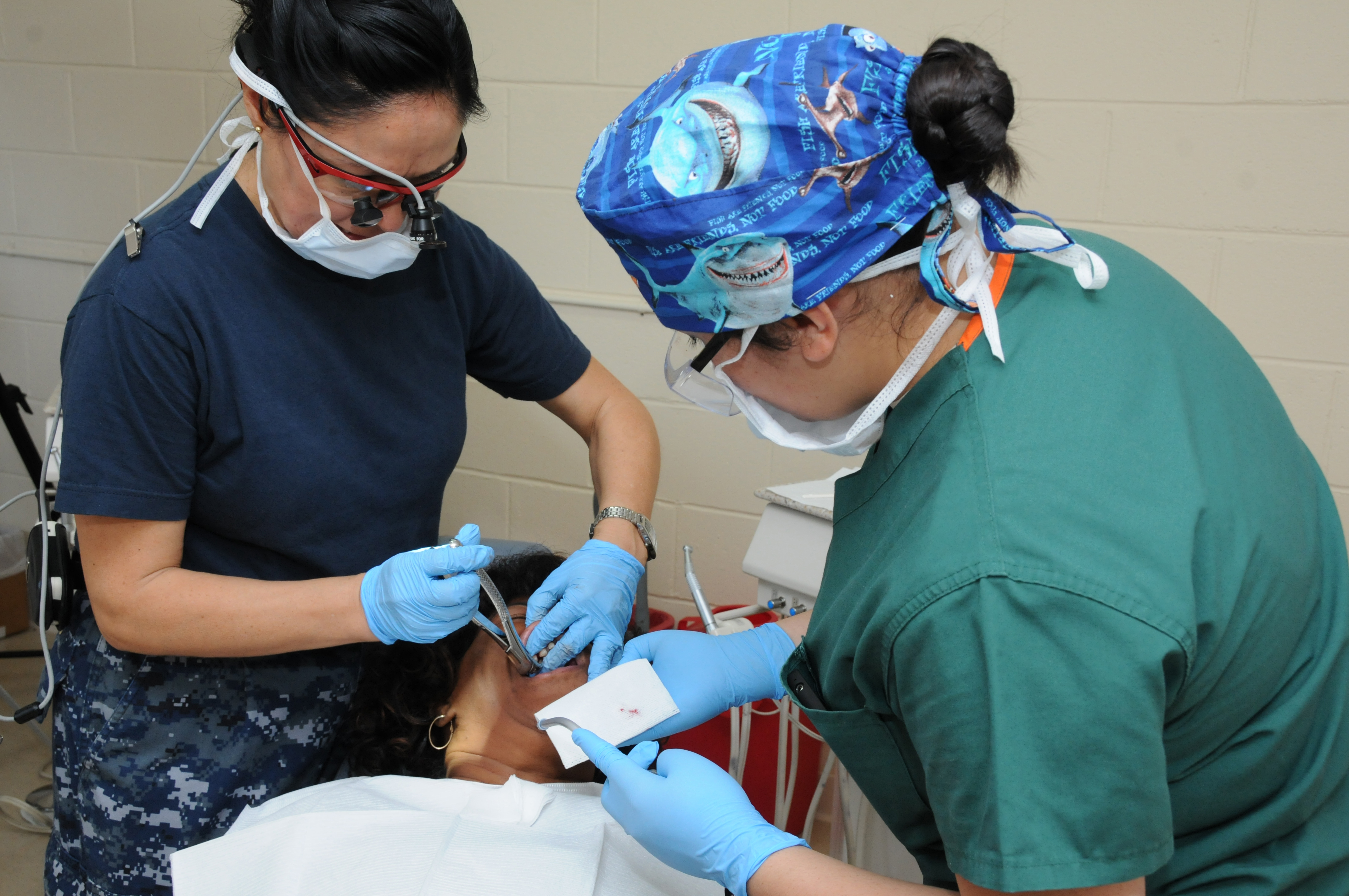 Two masked providers in scrubs perform a dental procedure on a patient in a clinic room.