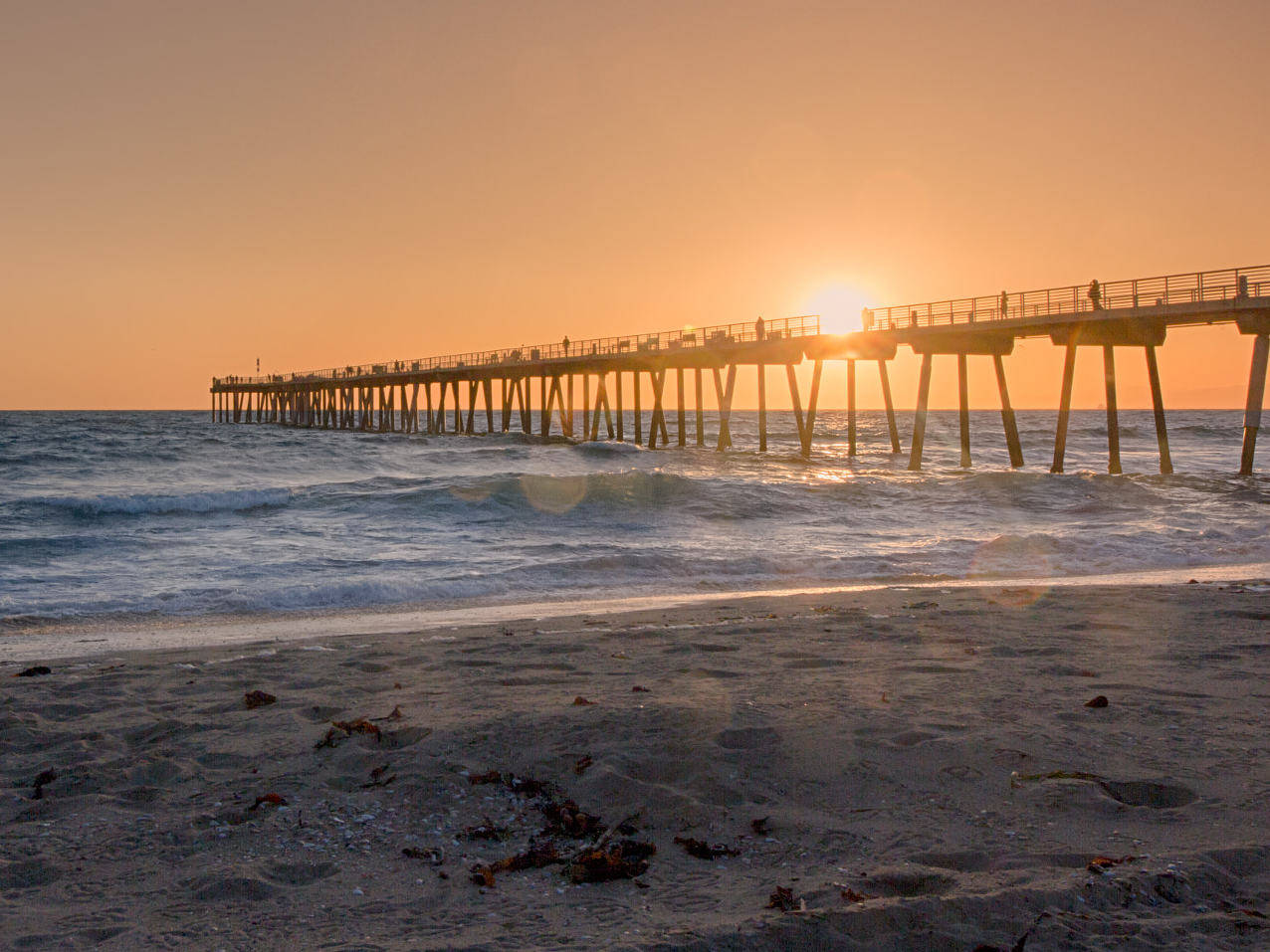 Wooden pier extending over the ocean at sunset with silhouetted figures and waves below.