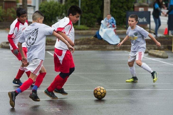 Youth soccer players in red and white uniforms competing for a ball on a wet paved court surface.