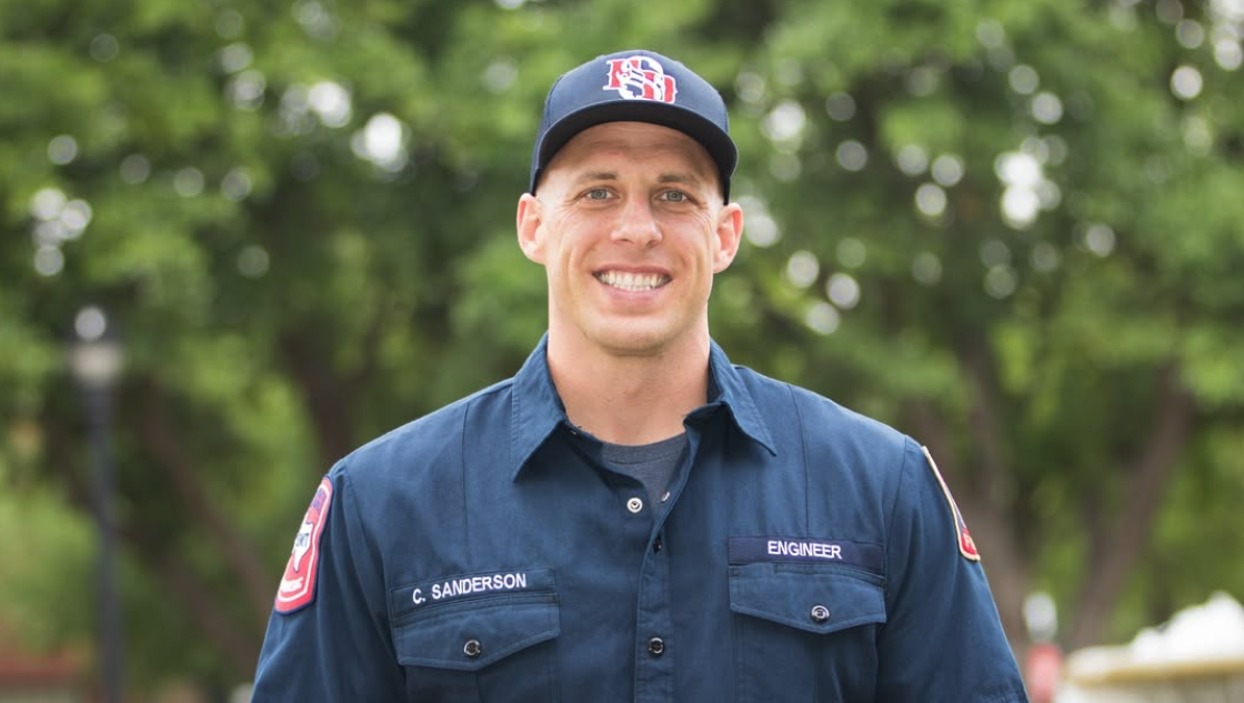 Southlake Fire Driver/Engineer Chase Sanderson smiles in his navy uniform and department cap outdoors.