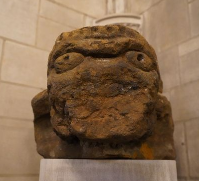 A weathered 15th-century carved stone grotesque known as "the Beast," featuring a face with deep-set eyes and pierced waterspout holes, displayed on a pedestal against a stone church interior.