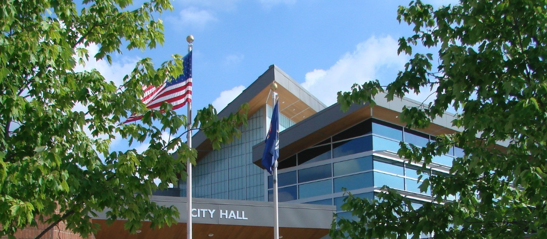 Farmington Hills City Hall exterior, framed by tree branches, with the American and Michigan state flags flying in front under a blue sky.