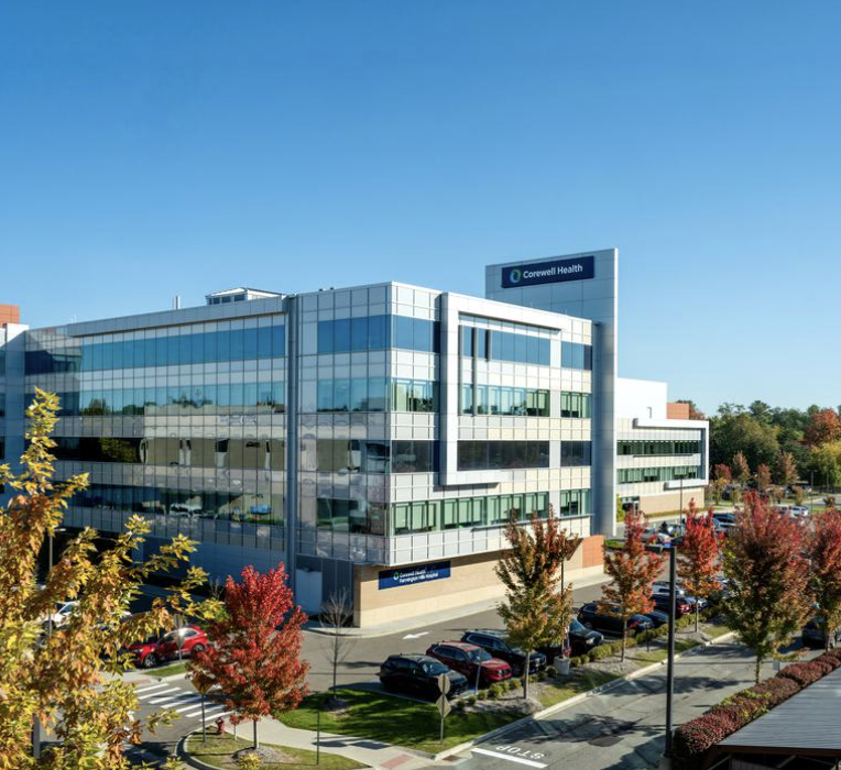 Exterior of Corewell Health Farmington Hills Hospital, a multi-story glass medical building with a landscaped parking lot in the foreground.