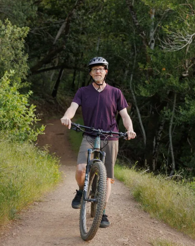 Craig Gleason wearing a bicycle helmet and holding a mountain bike on a wooded dirt trail.