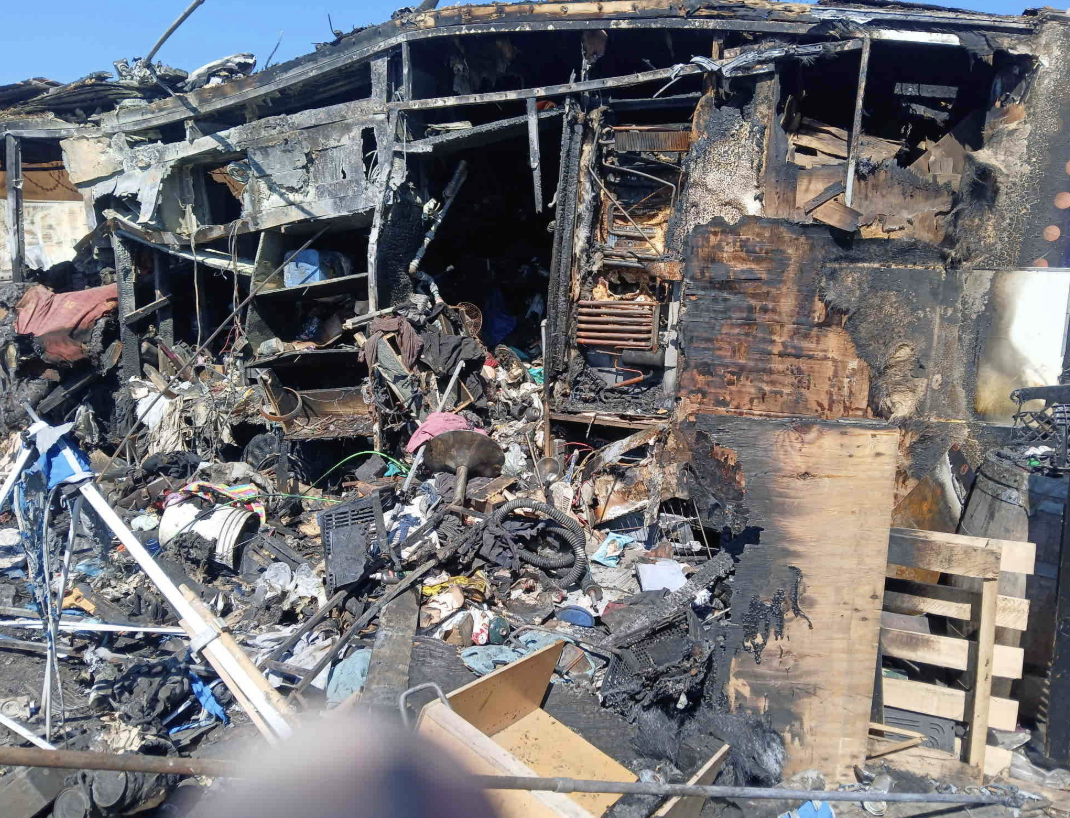 The charred, collapsed interior of a burned trailer, with blackened walls, melted fixtures and scattered scorched belongings
