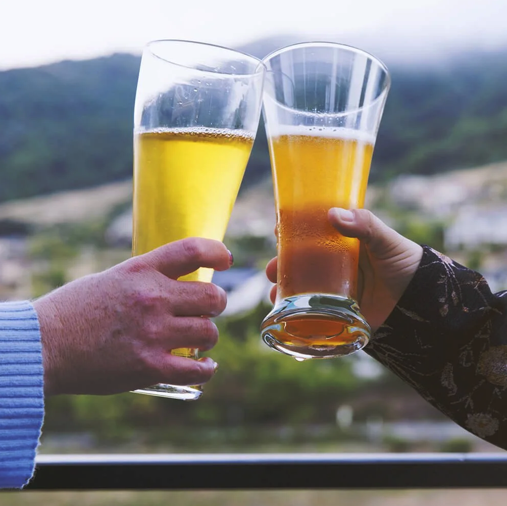 Two hands clink pint glasses of beer on an outdoor patio overlooking green hills, part of CreekHaus's planned beer garden in Orinda.