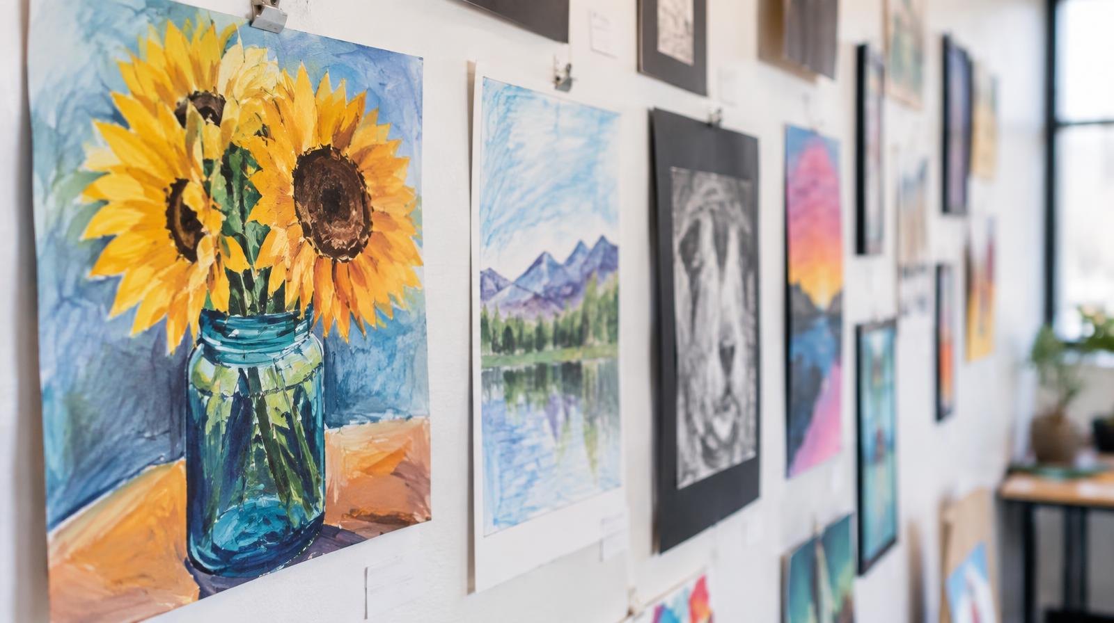 Colorful paintings and drawings arranged on a bright white gallery wall.