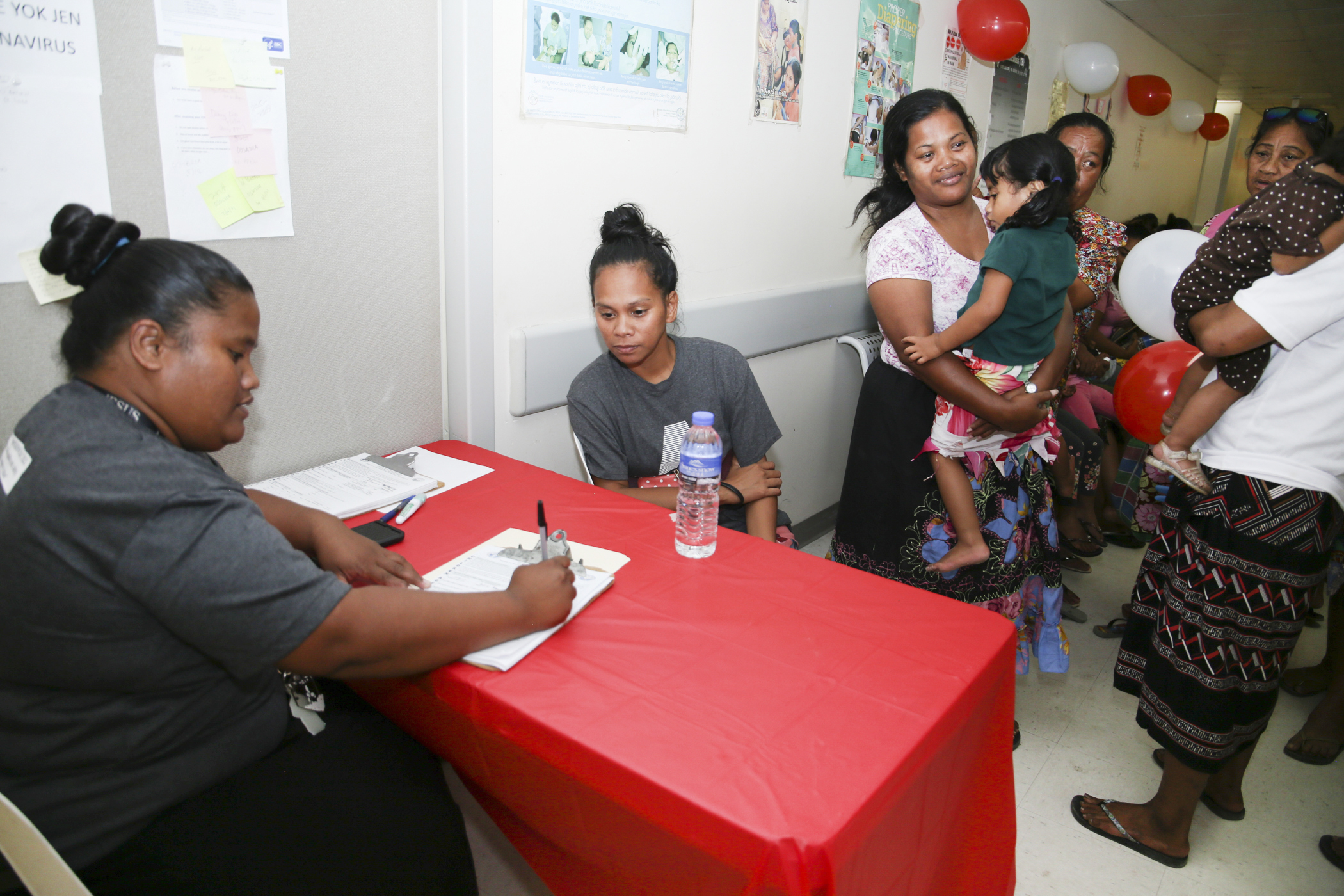 Parents and children queue at a vaccination clinic registration table staffed by two workers.