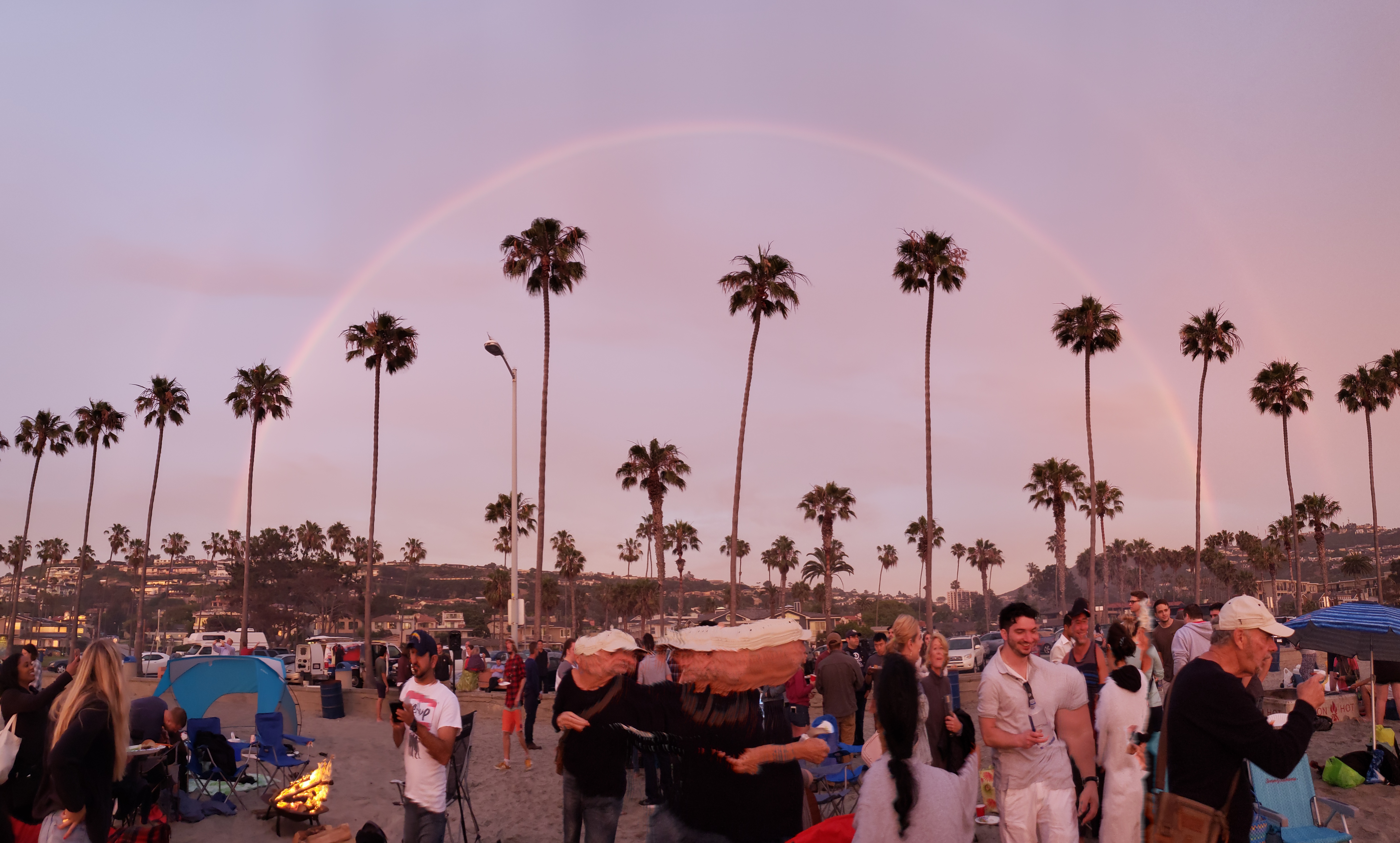 Large crowd on a palm-lined beach at sunset beneath a double rainbow.