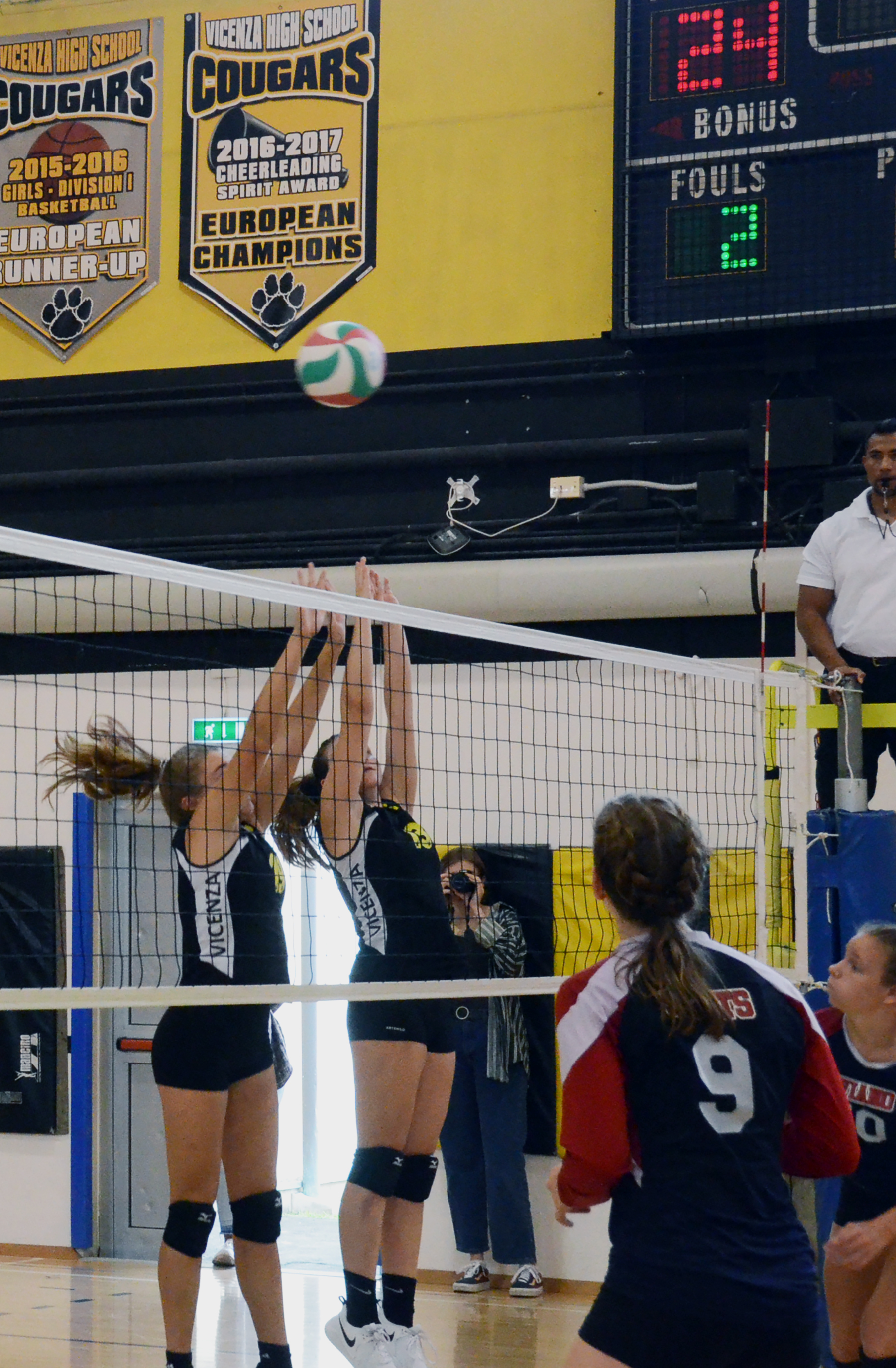 Two female volleyball players jump to block at the net inside a gymnasium.