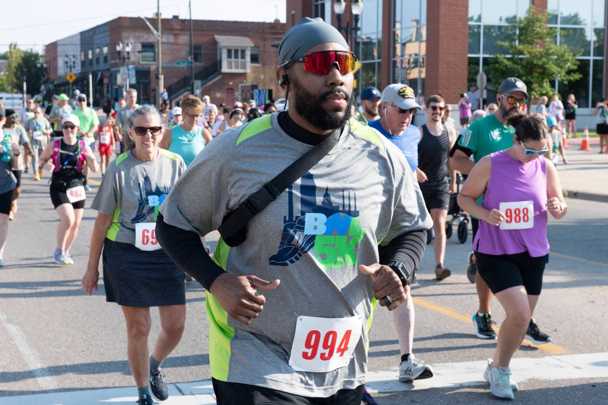 Runners wearing numbered race bibs jog along an urban street on a sunny day.