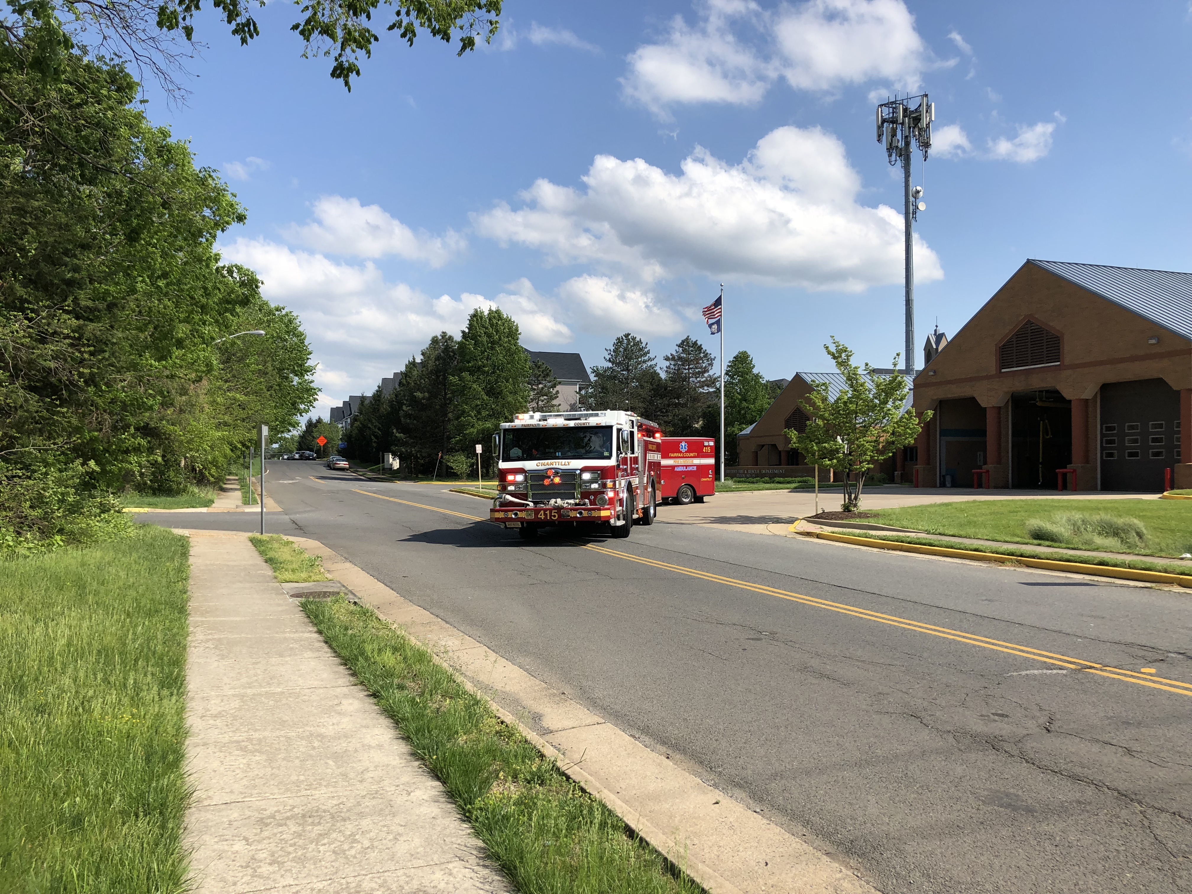 A red and white Fairfax County Fire and Rescue fire truck on a road in front of a brick fire station.