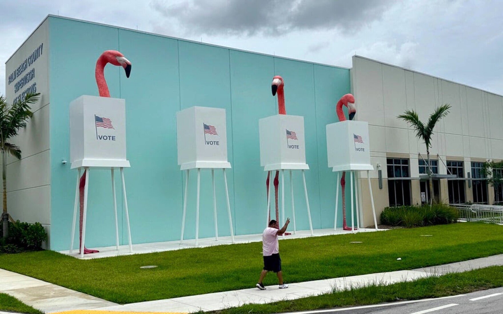 Three large voting booth sculptures with American flags and VOTE signs flanked by pink flamingo statues outside a turquoise building.