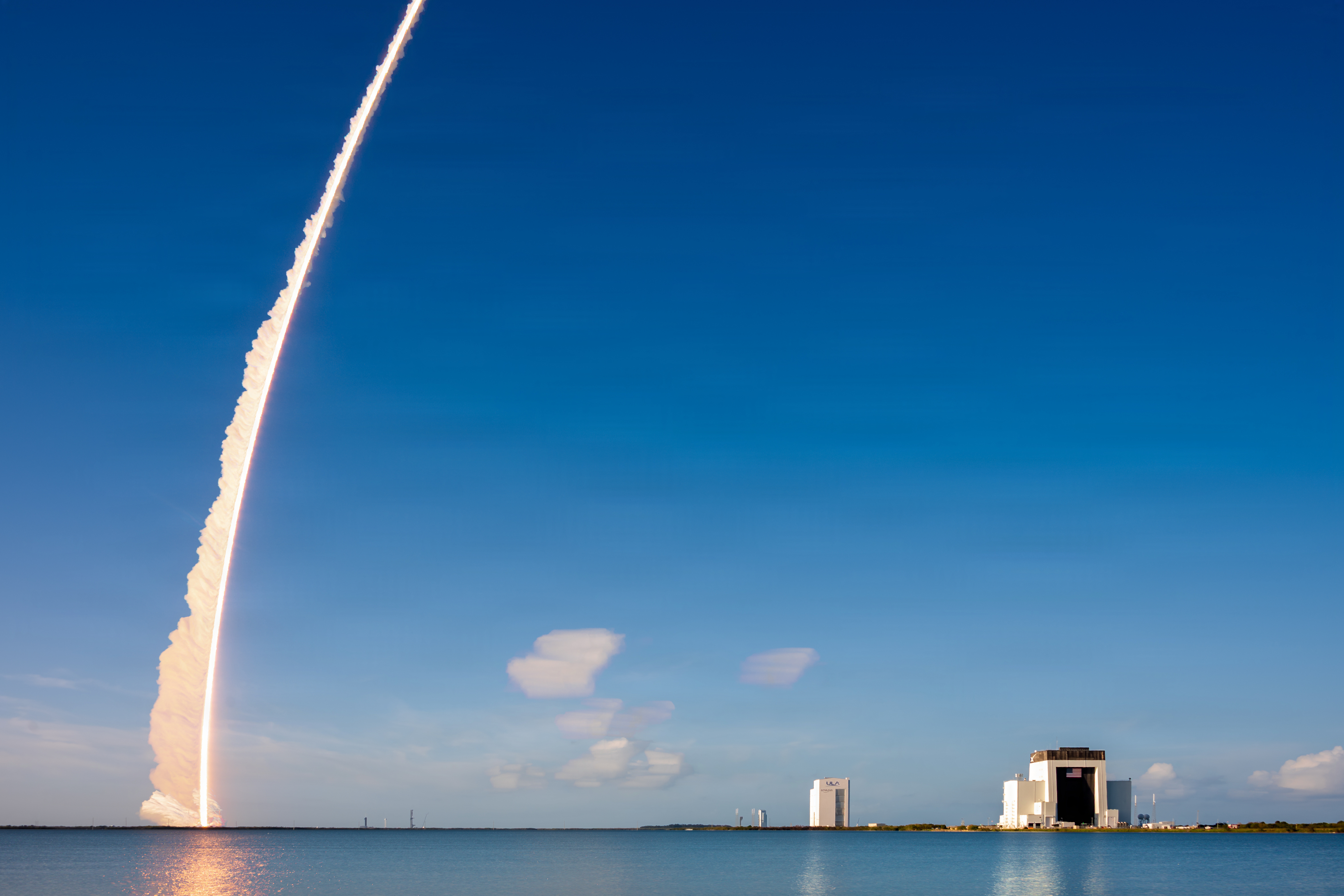 Rocket ascending from a launch pad with a bright exhaust trail reflected in water below.