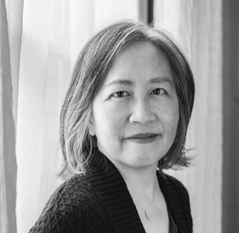 Black-and-white portrait of a woman with shoulder-length hair wearing a dark cardigan, standing near a window.