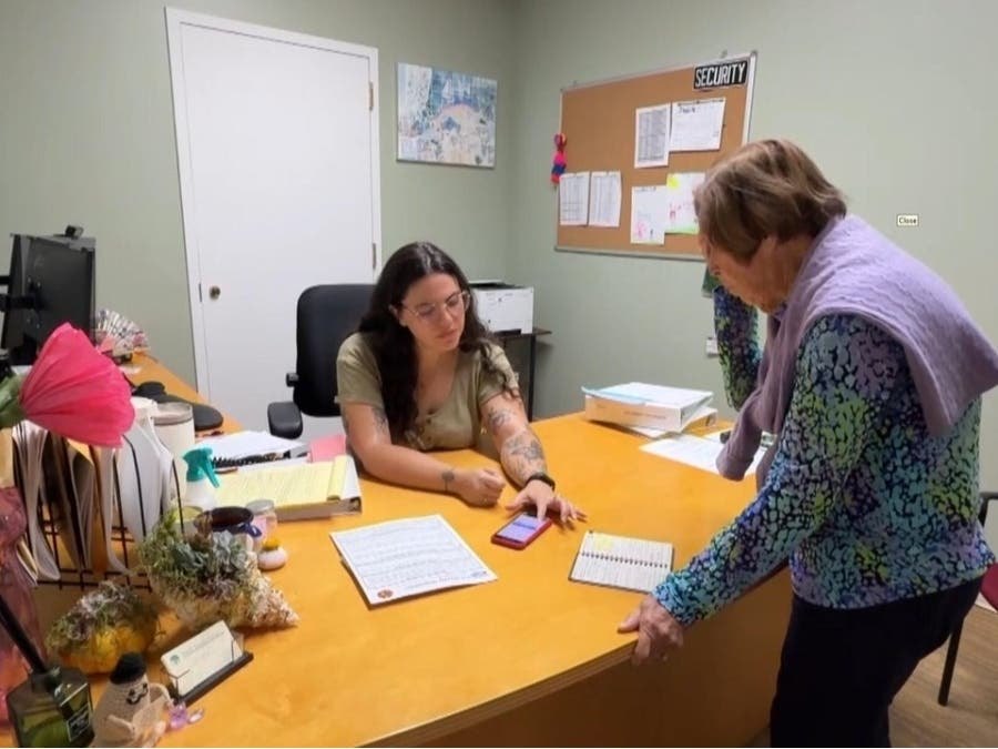 Two women at an office desk, one seated showing something on a phone to the other standing nearby.