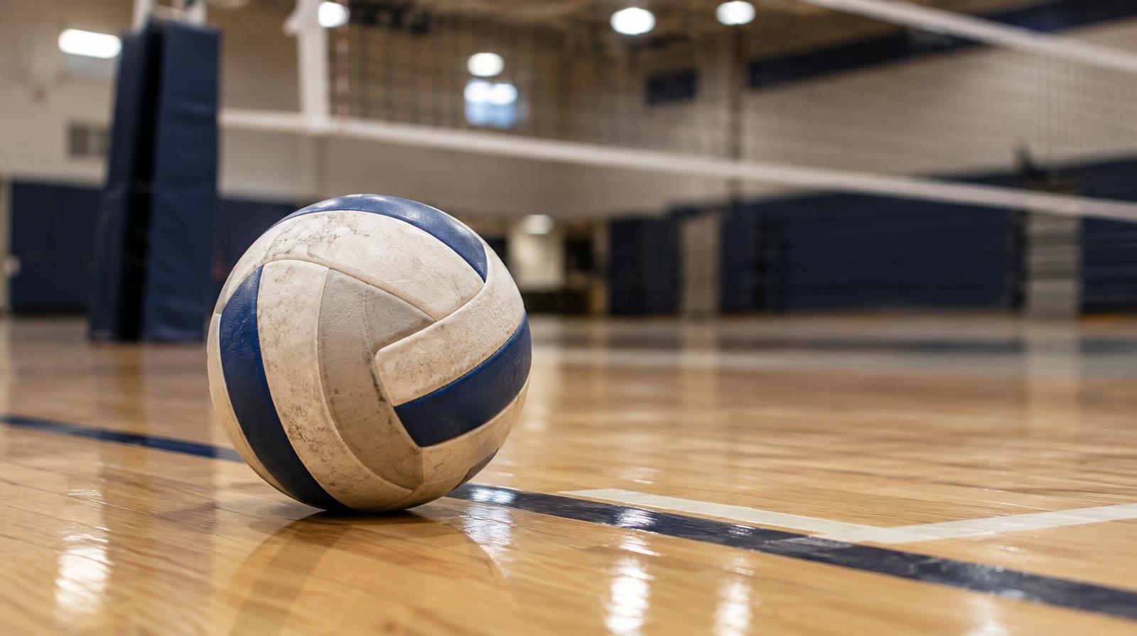 A volleyball resting on a hardwood gym floor near the net under bright lights.