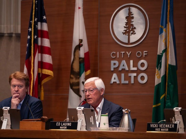 Two men seated at the Palo Alto City Council dais with the city seal and flags behind them.