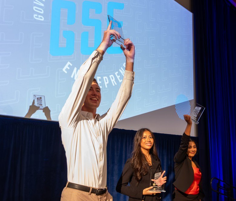 Three young people on a stage holding glass trophies and celebrating in front of a GSE banner.