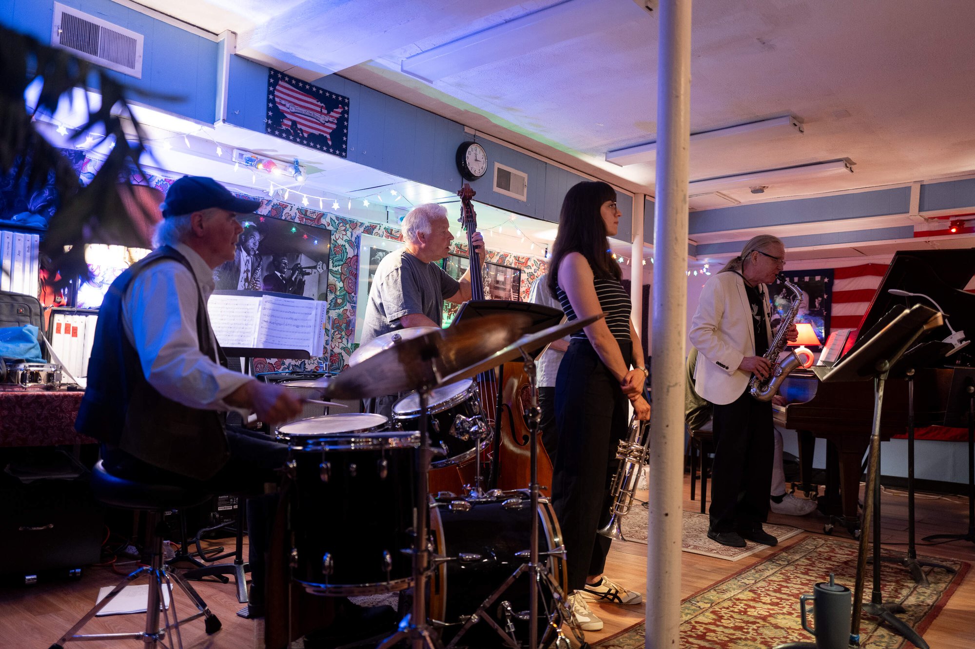 A jazz band performs on a small stage in a dimly lit basement venue with American flags on the walls.