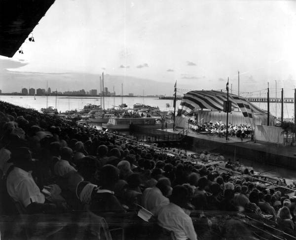 Black-and-white photo of a packed stadium crowd watching a waterfront concert with boats and a city skyline in the background
