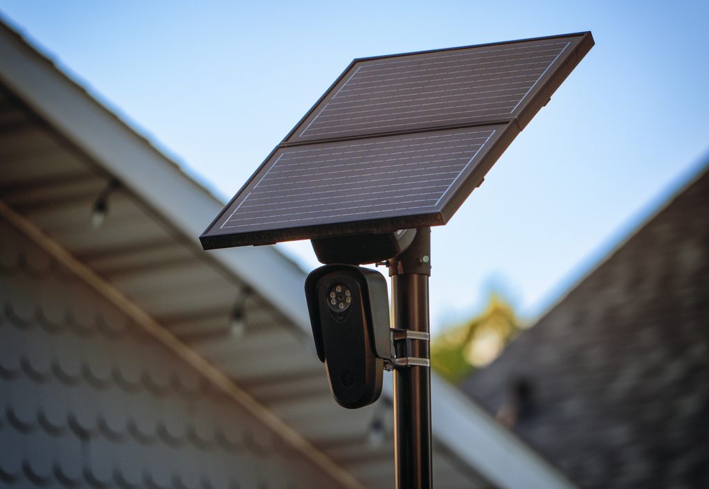 A Flock Safety automatic license plate recognition camera mounted on a pole with a solar panel, with a residential roofline and blue sky in the background.