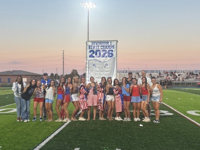 Girls lacrosse team poses on a football field in front of a state championship banner.