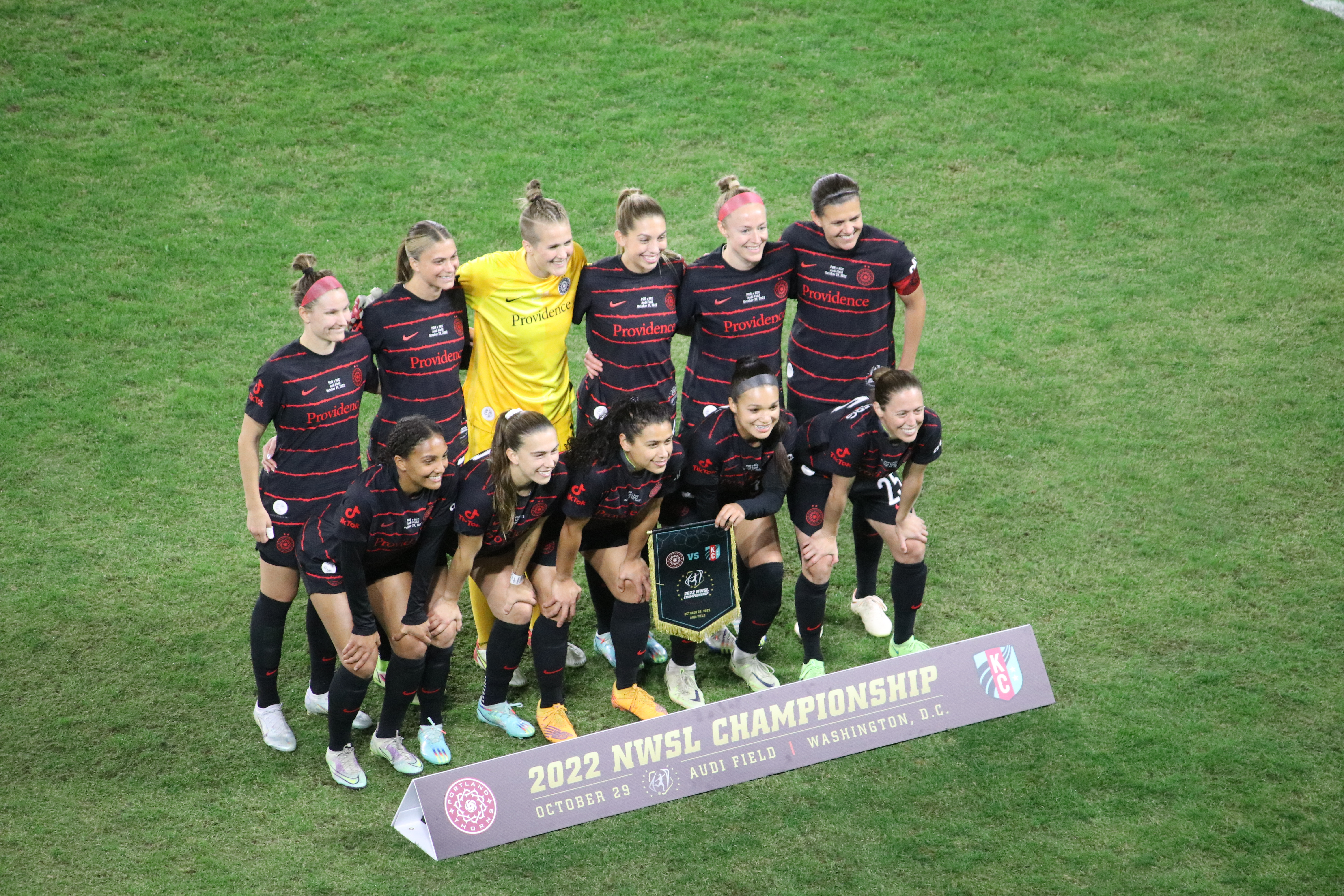 Women's soccer players in black and red uniforms pose in two rows on a grass field.