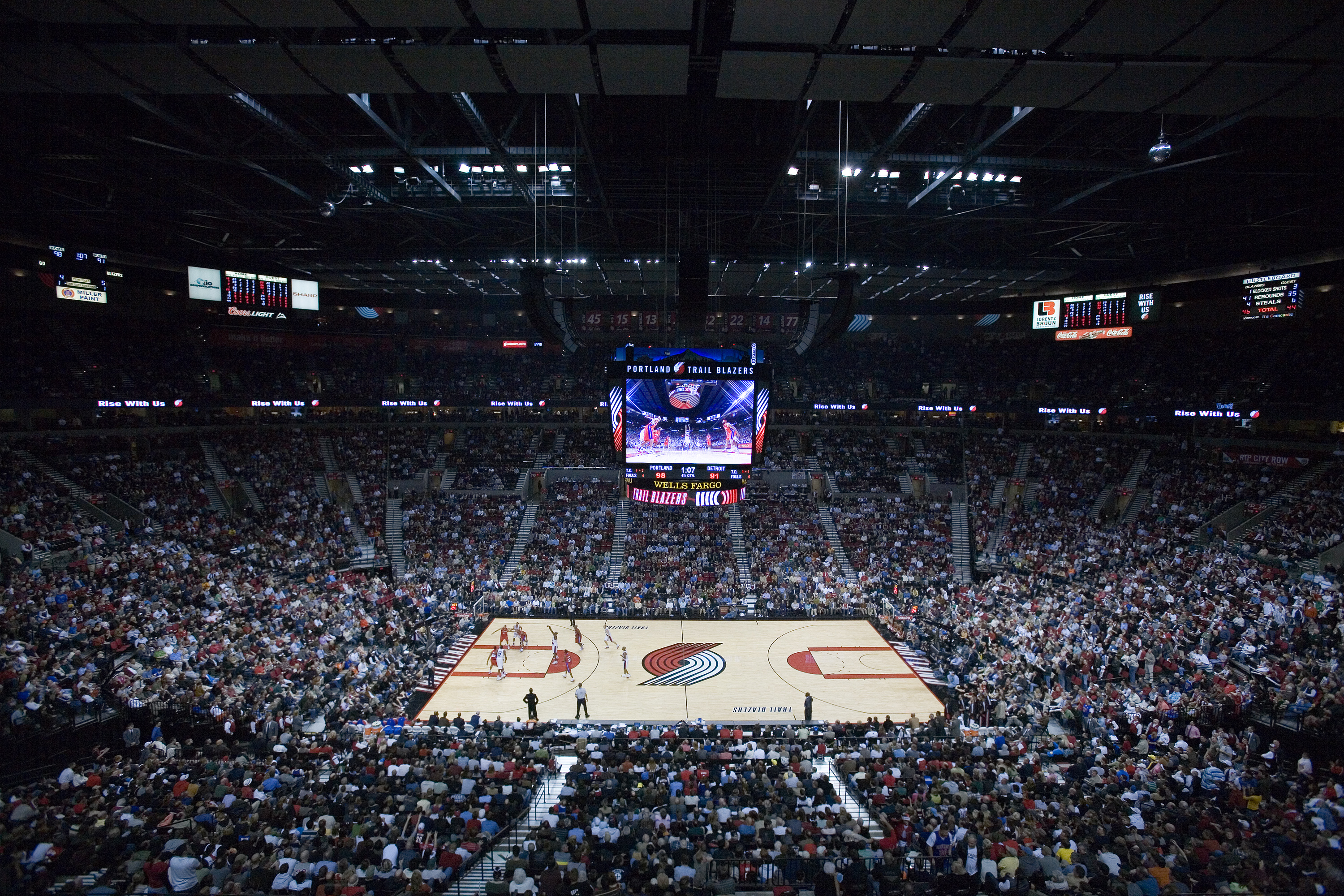 Full basketball arena interior packed with fans, court and jumbotron visible from upper level.
