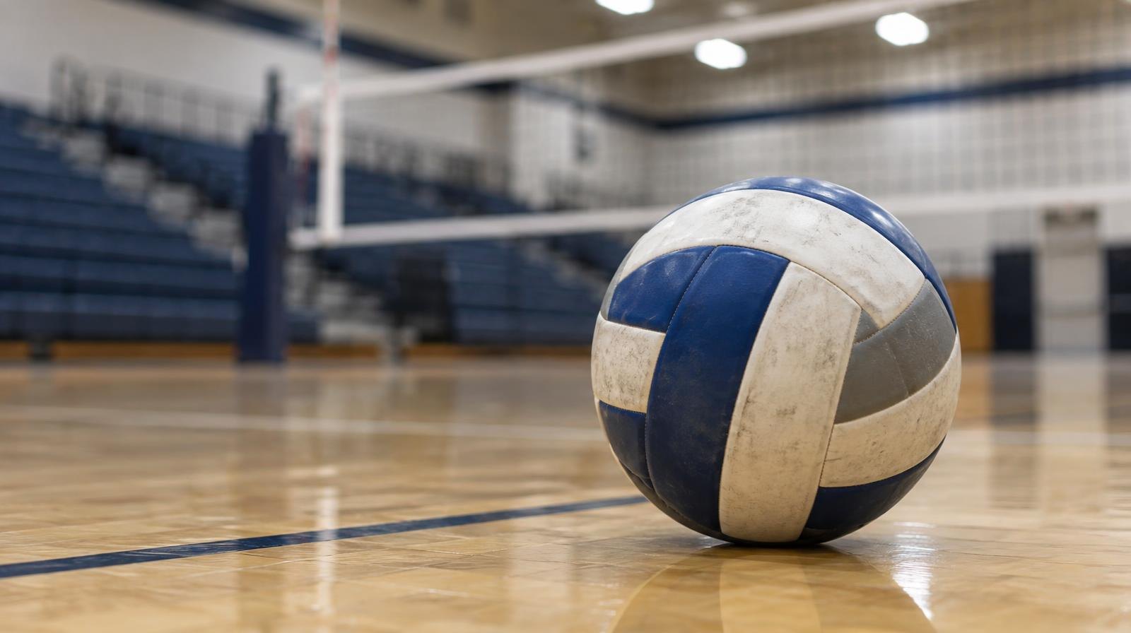 A volleyball resting on a hardwood gym floor near the net with bleachers in the background.