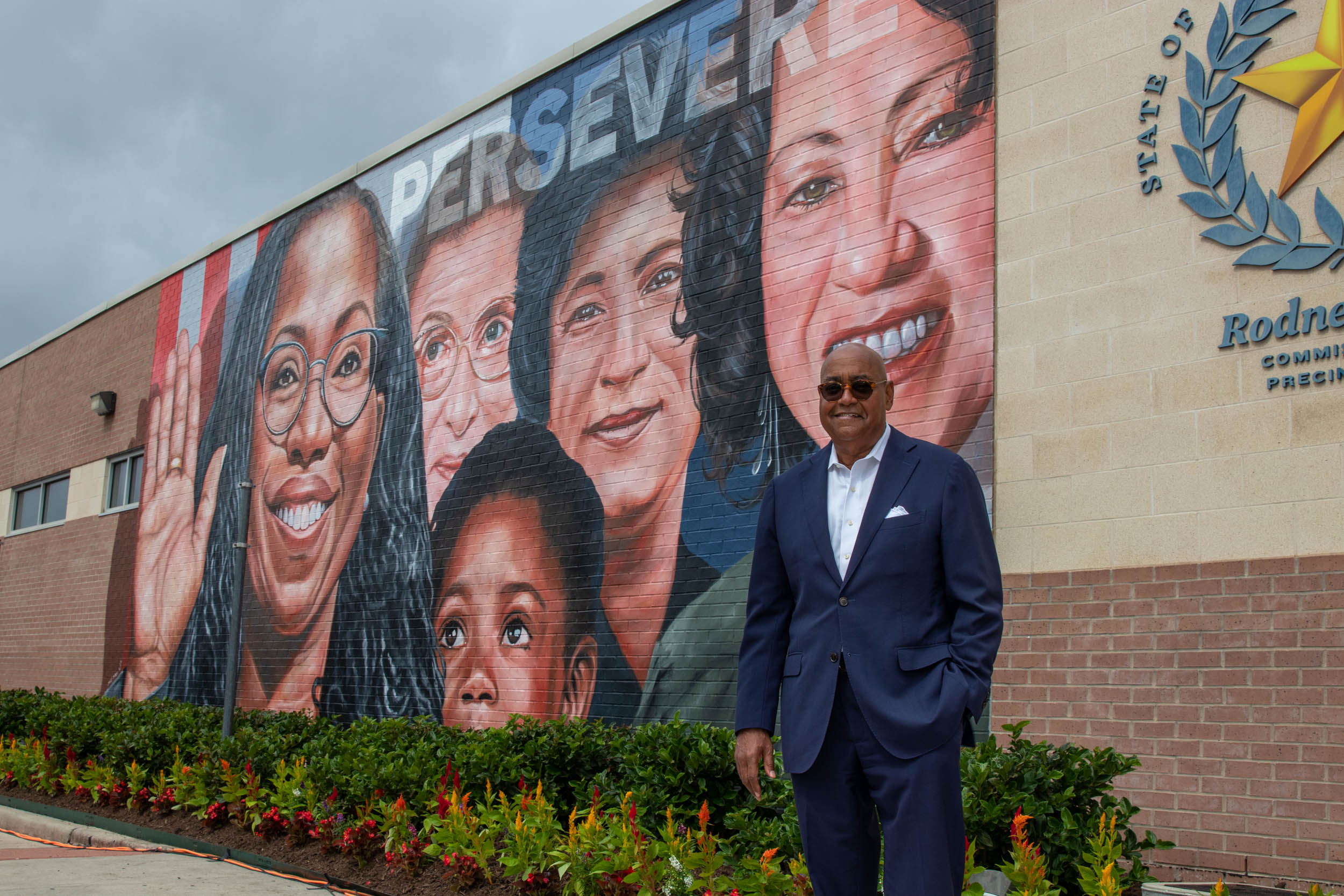 Man in a blue suit standing in front of a large mural on a brick building with the word 'PERSEVERE' and portraits of women.