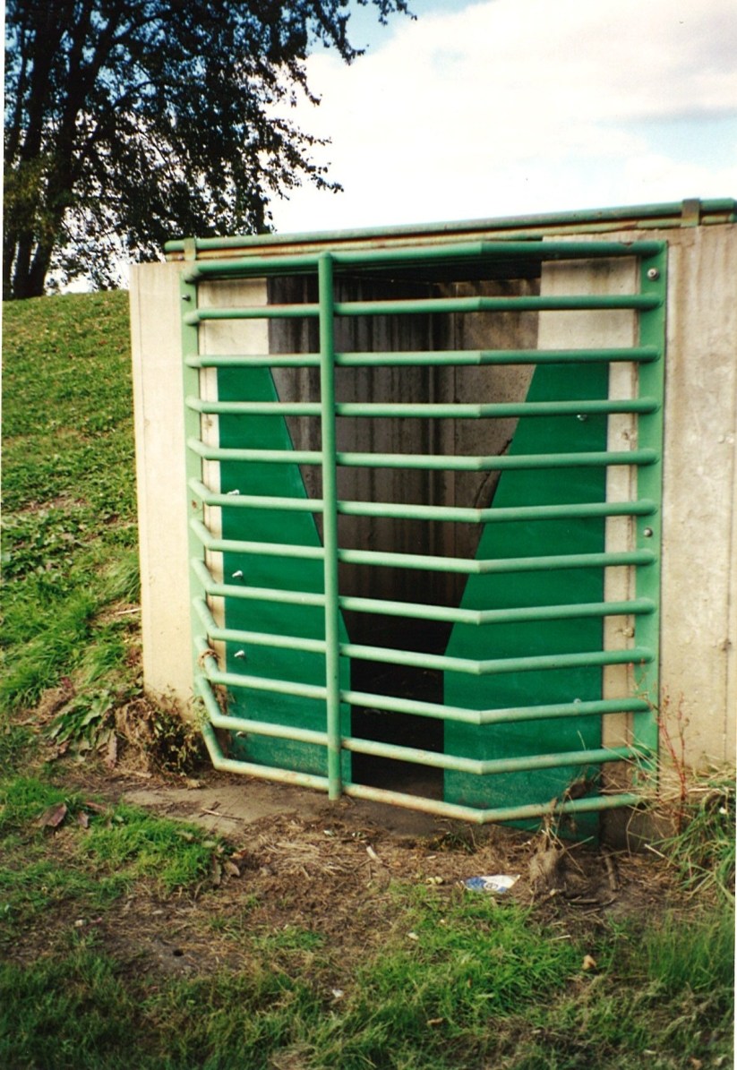 A green steel-grated outlet structure bolted onto a concrete stormwater detention basin embankment.