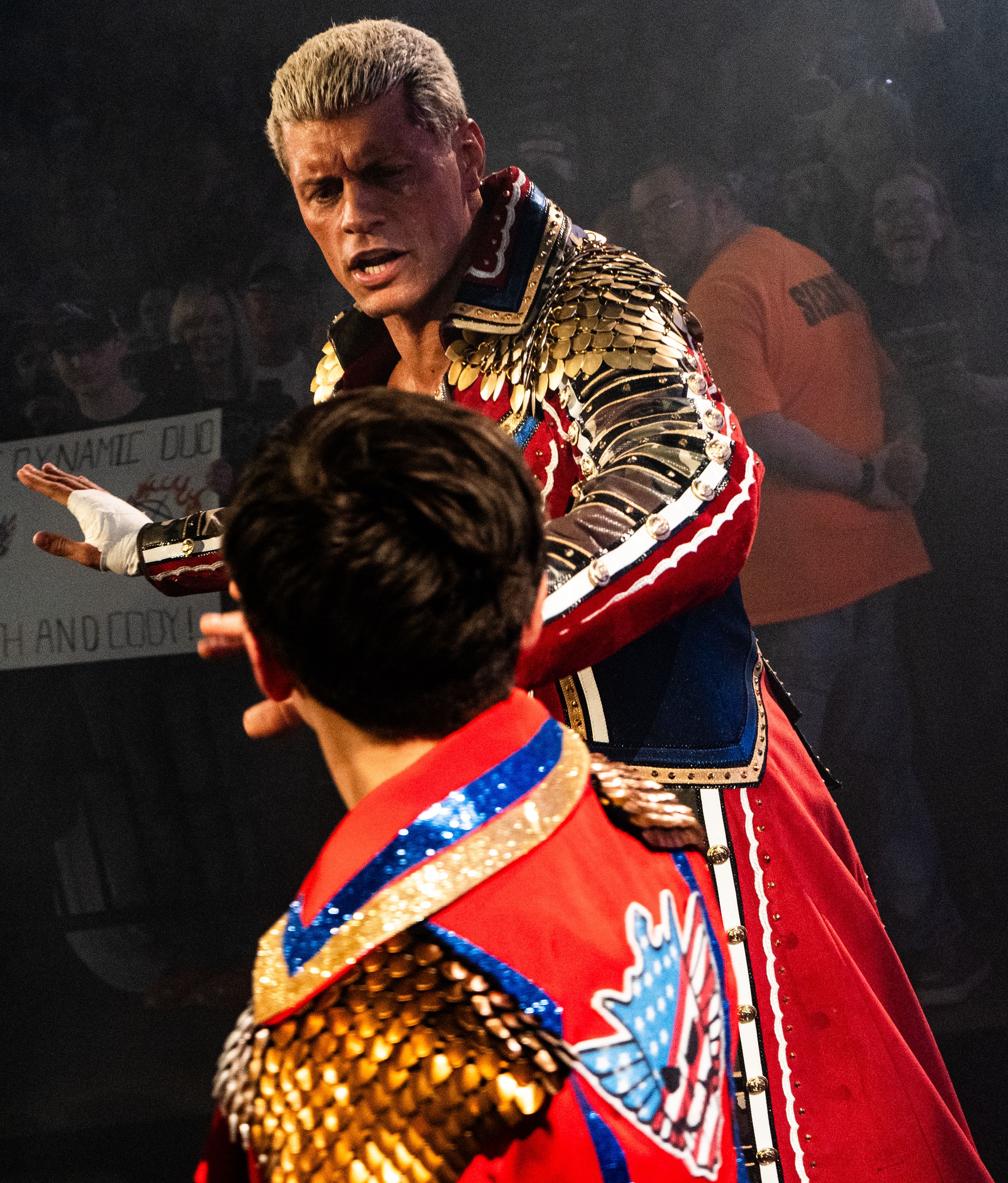 Adult wrestler in sequined ring gear leans toward a child in matching costume at an arena.