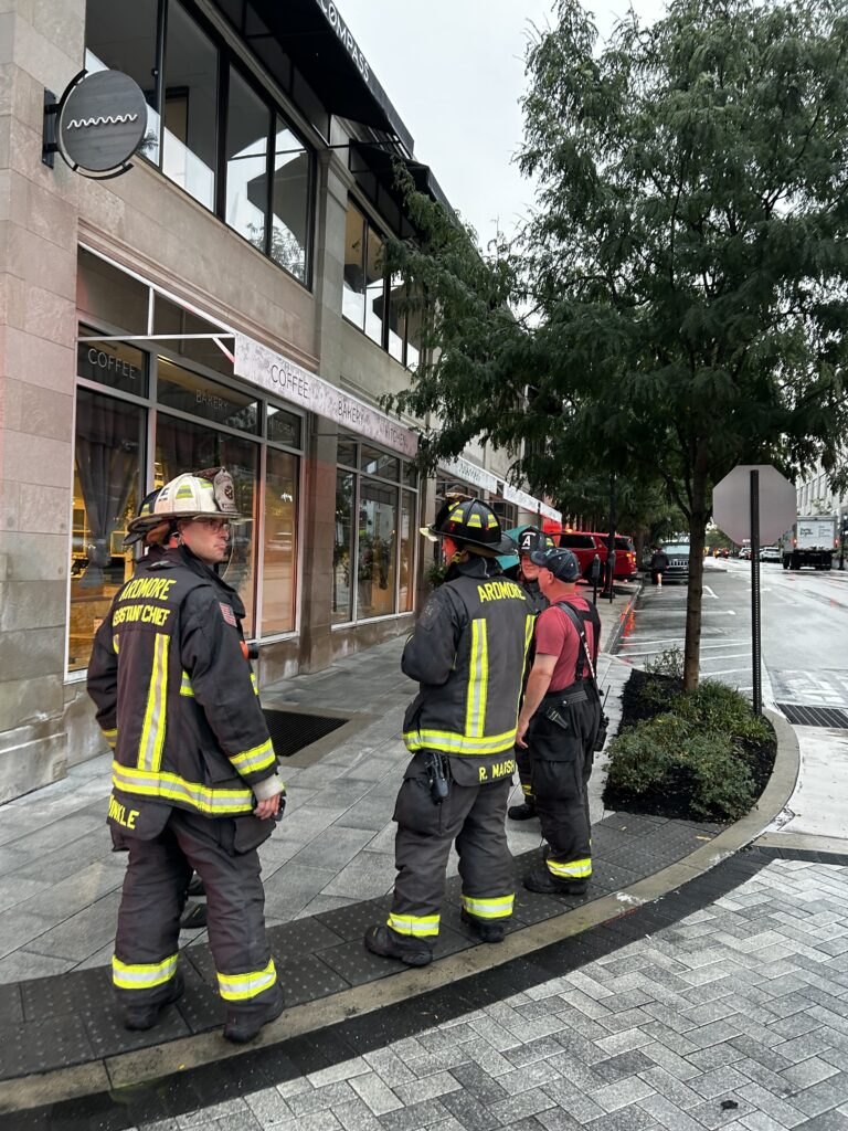 Three firefighters in black turnout gear labeled 'Ardmore' stand on a sidewalk outside a storefront.