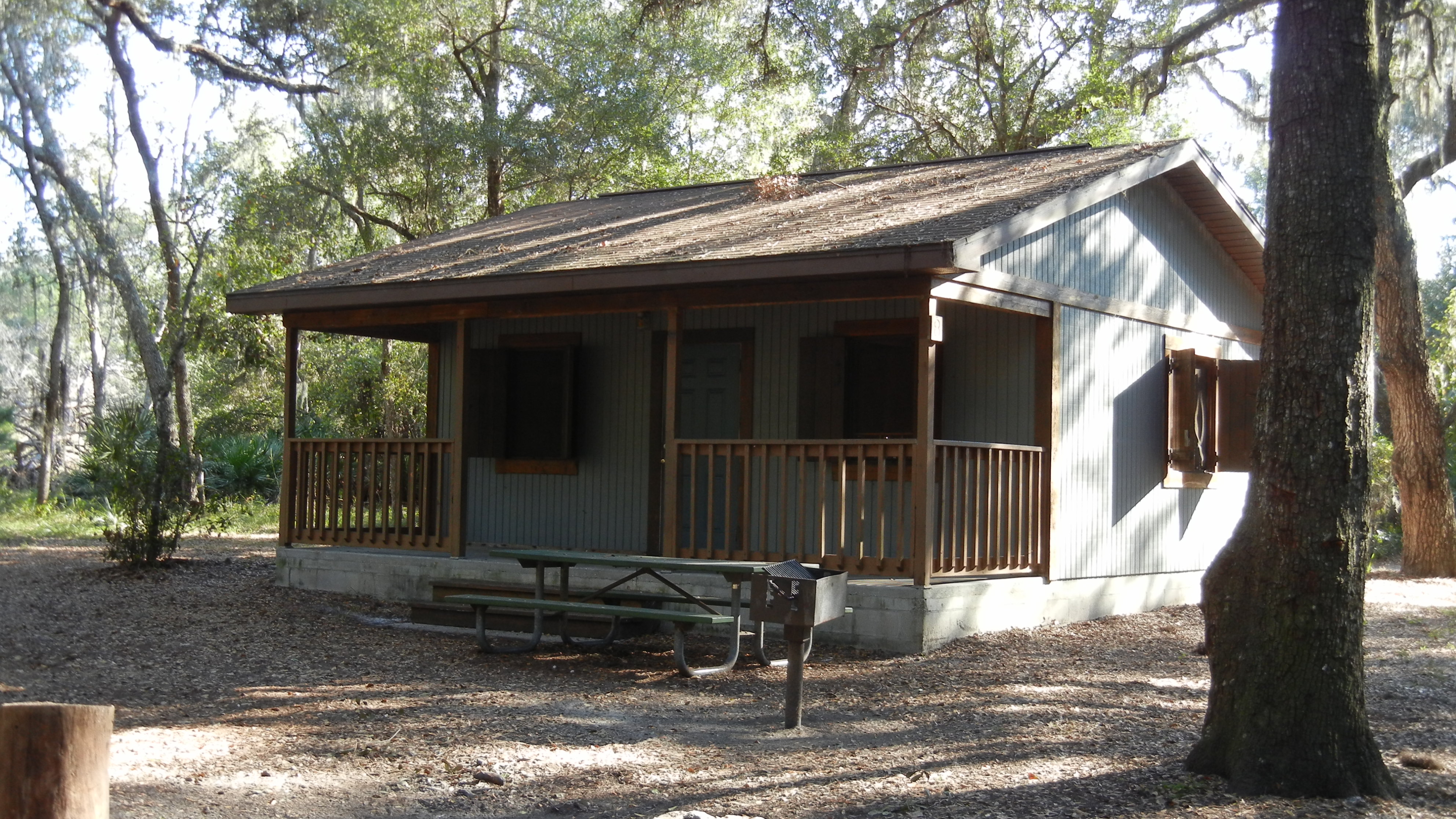 A small blue cabin with a covered porch surrounded by oak trees at a wilderness park.