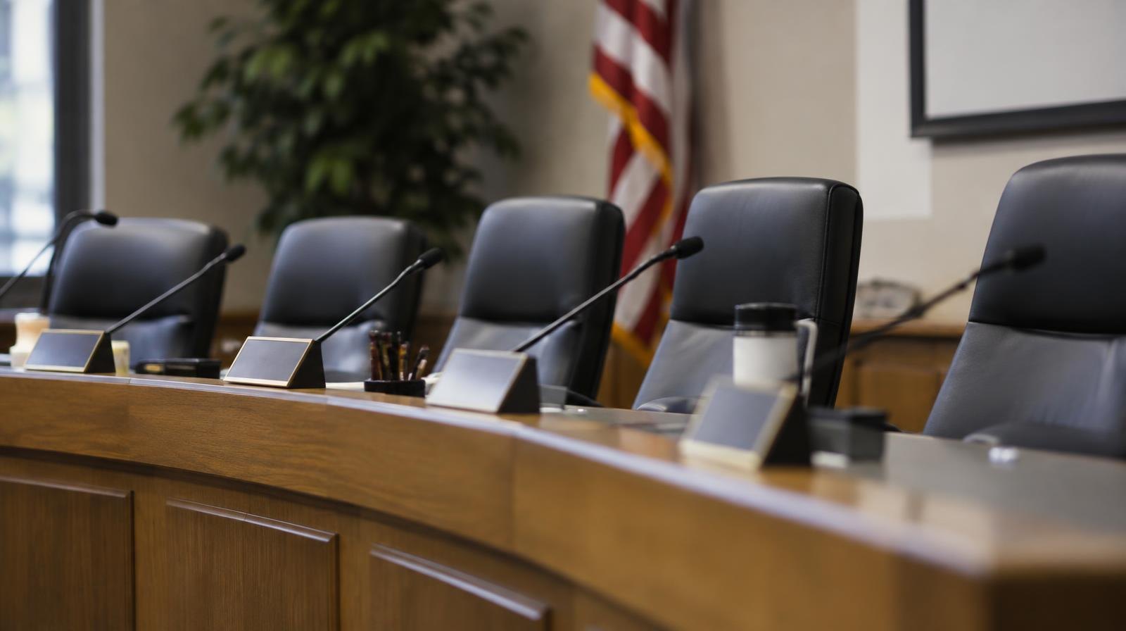 Empty chairs behind a long table at a school board meeting dais.