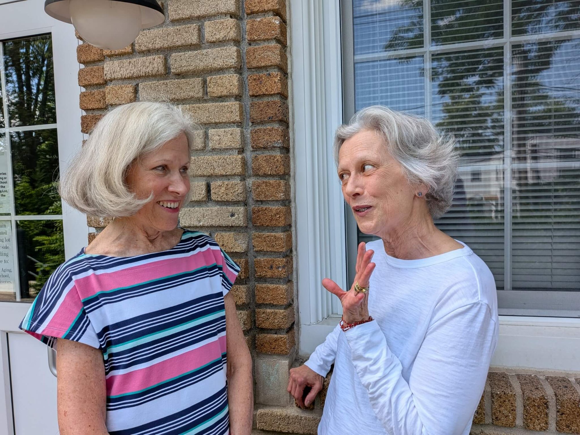 Two older women with gray hair stand outside a brick building, talking.